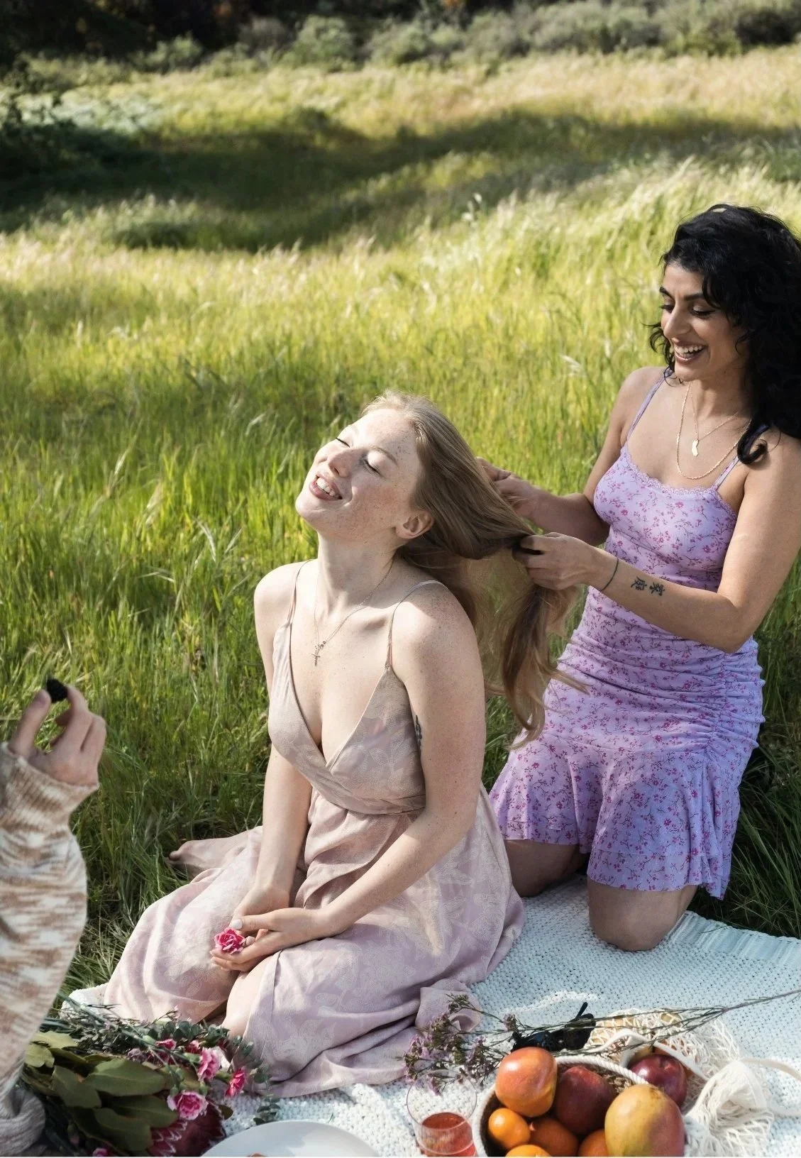 Two women enjoying a picnic outdoors in a grassy field; one woman with red hair sitting on a blanket, the other with black hair kneeling behind her, holding and playing with her hair while smiling.