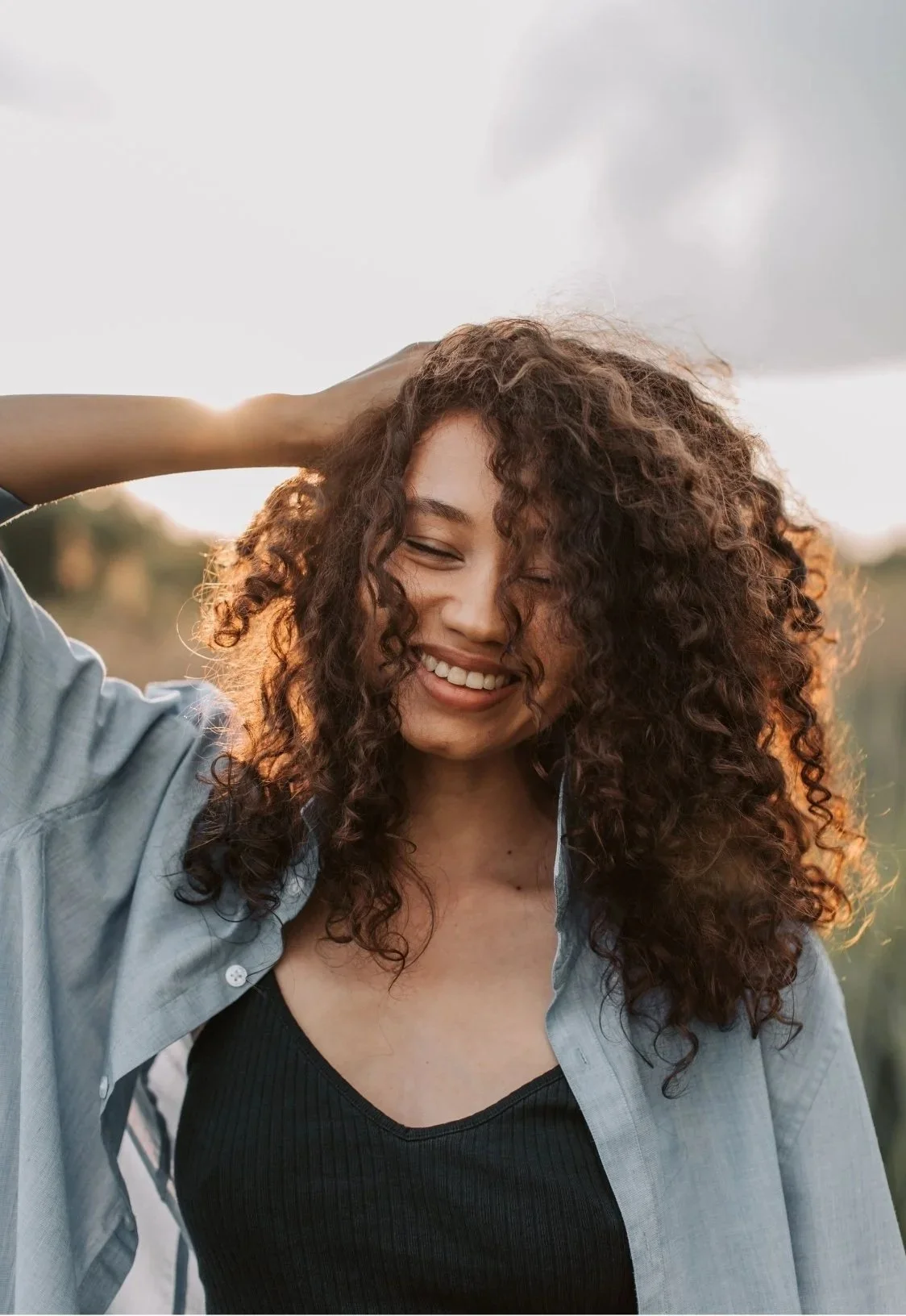 A woman with curly hair smiling with her eyes closed, wearing a denim shirt over a black top, outdoors during sunset.