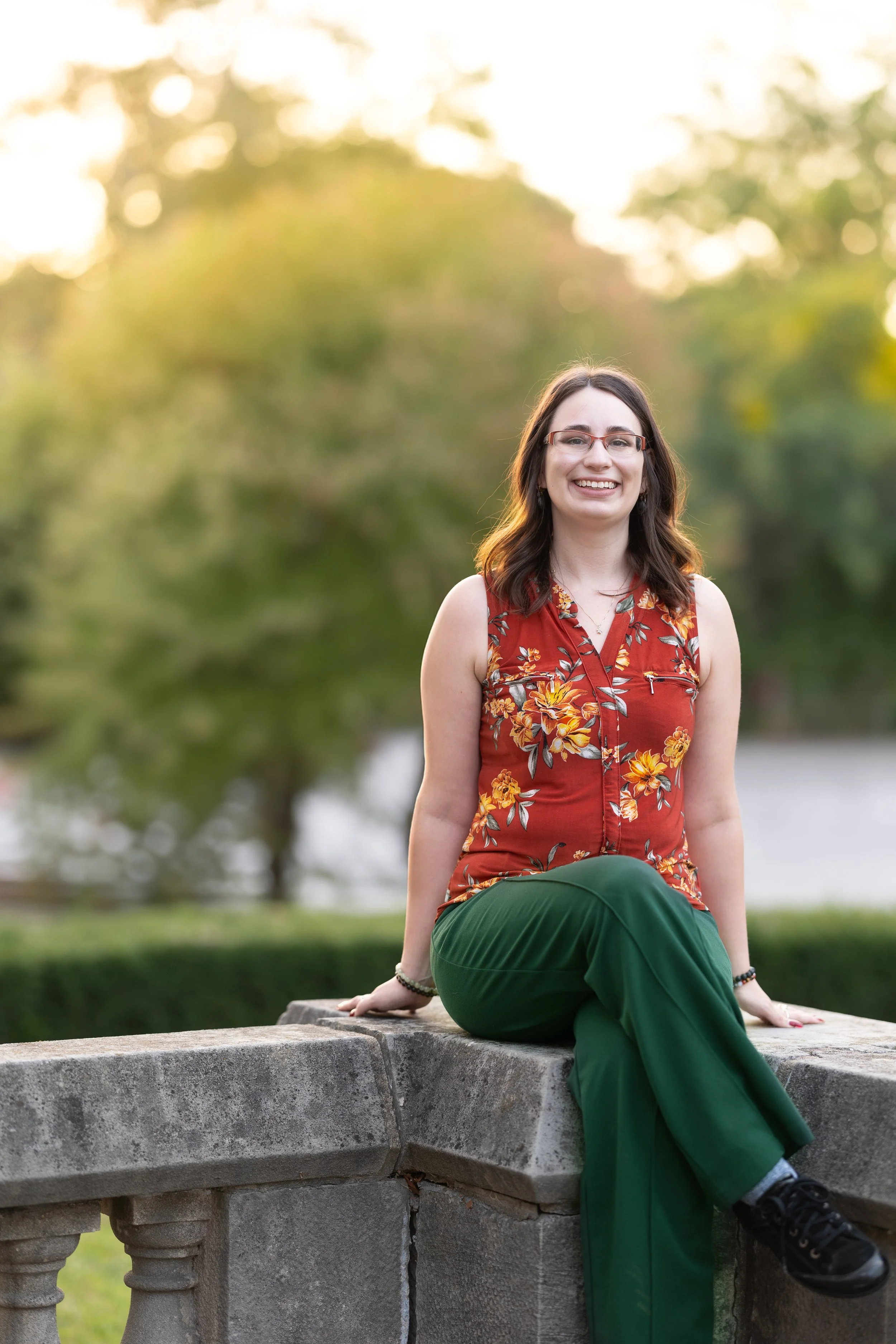 Hillary Bowdridge, Psychotherapist in Richmond Hill, ON, with glasses, wearing a sleeveless floral top and green pants, sitting on a stone ledge outdoors with trees and sunlight in the background.