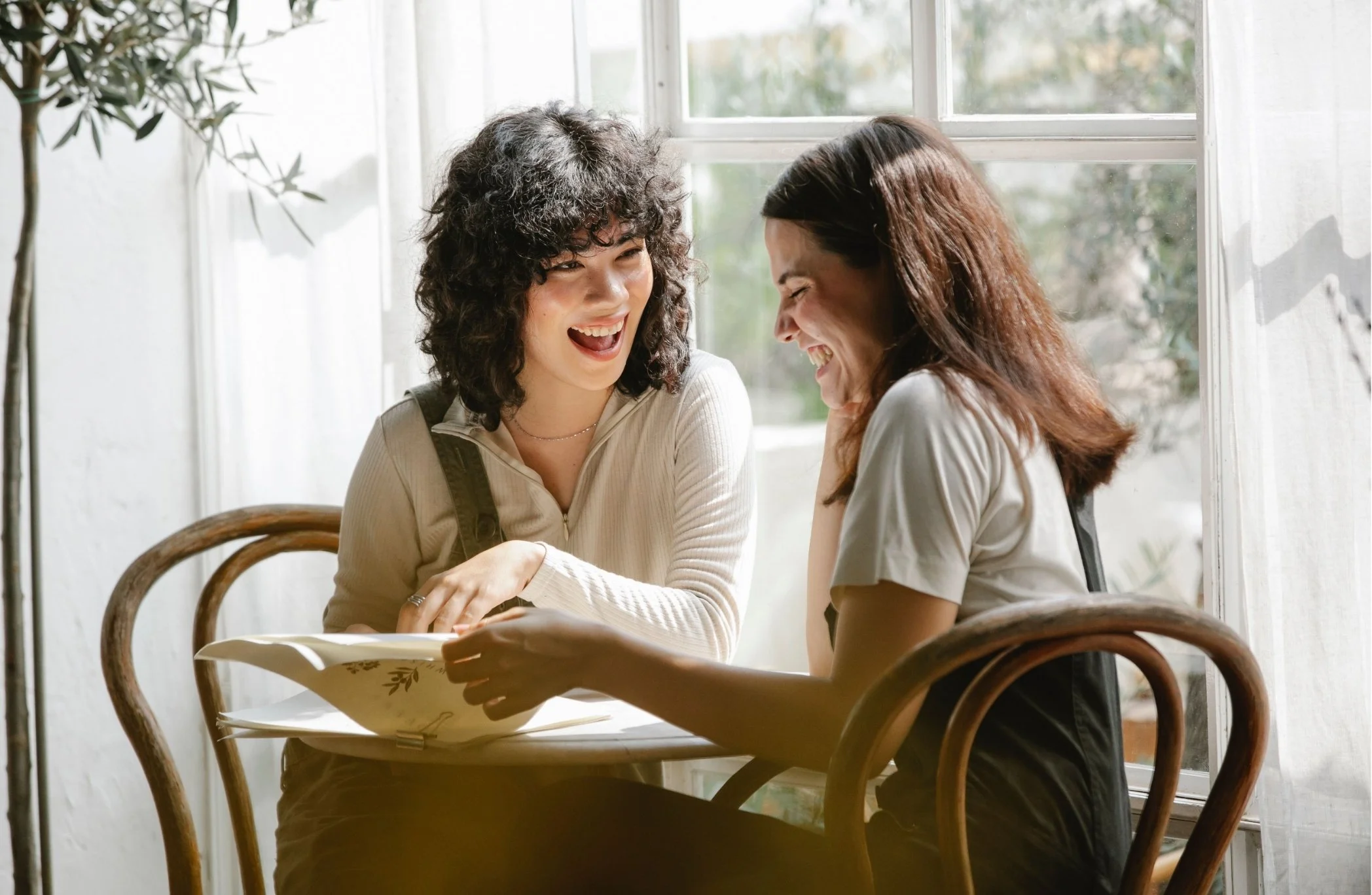 Two women are sitting at a wooden table near a large window, laughing and enjoying a conversation.