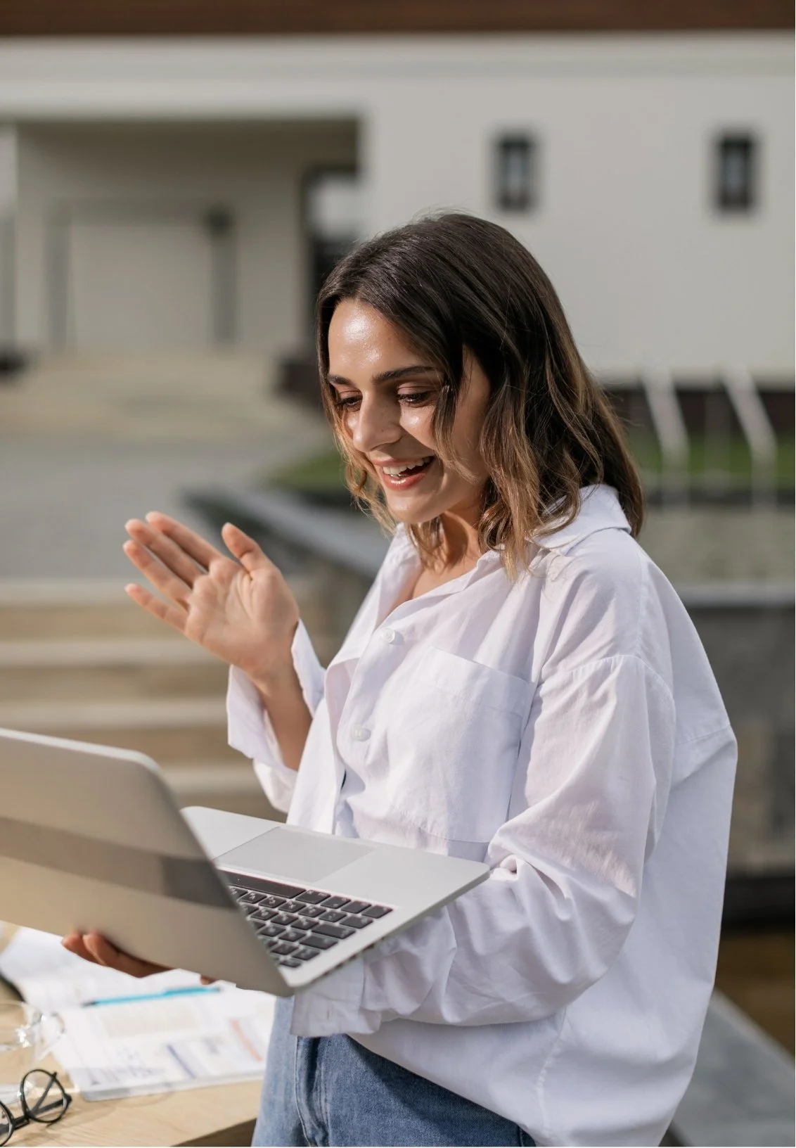 A woman in a white shirt holding a laptop and smiling outdoors.