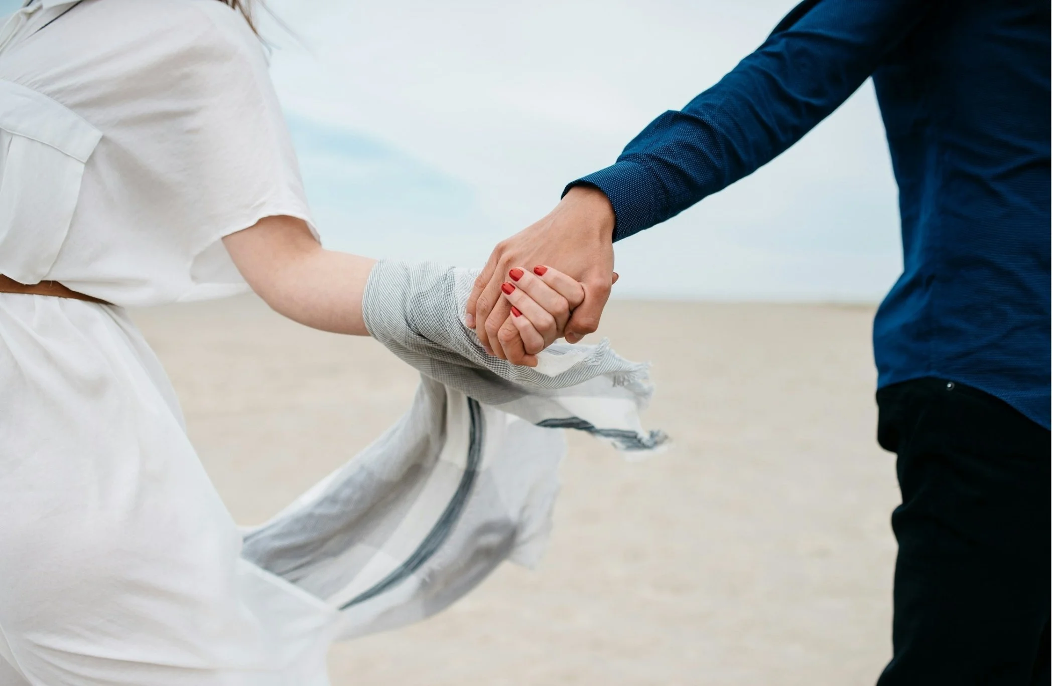 Close-up of a couple holding hands, with the woman wearing red nail polish and a white dress, and the man wearing a blue shirt, on a beach during daytime.