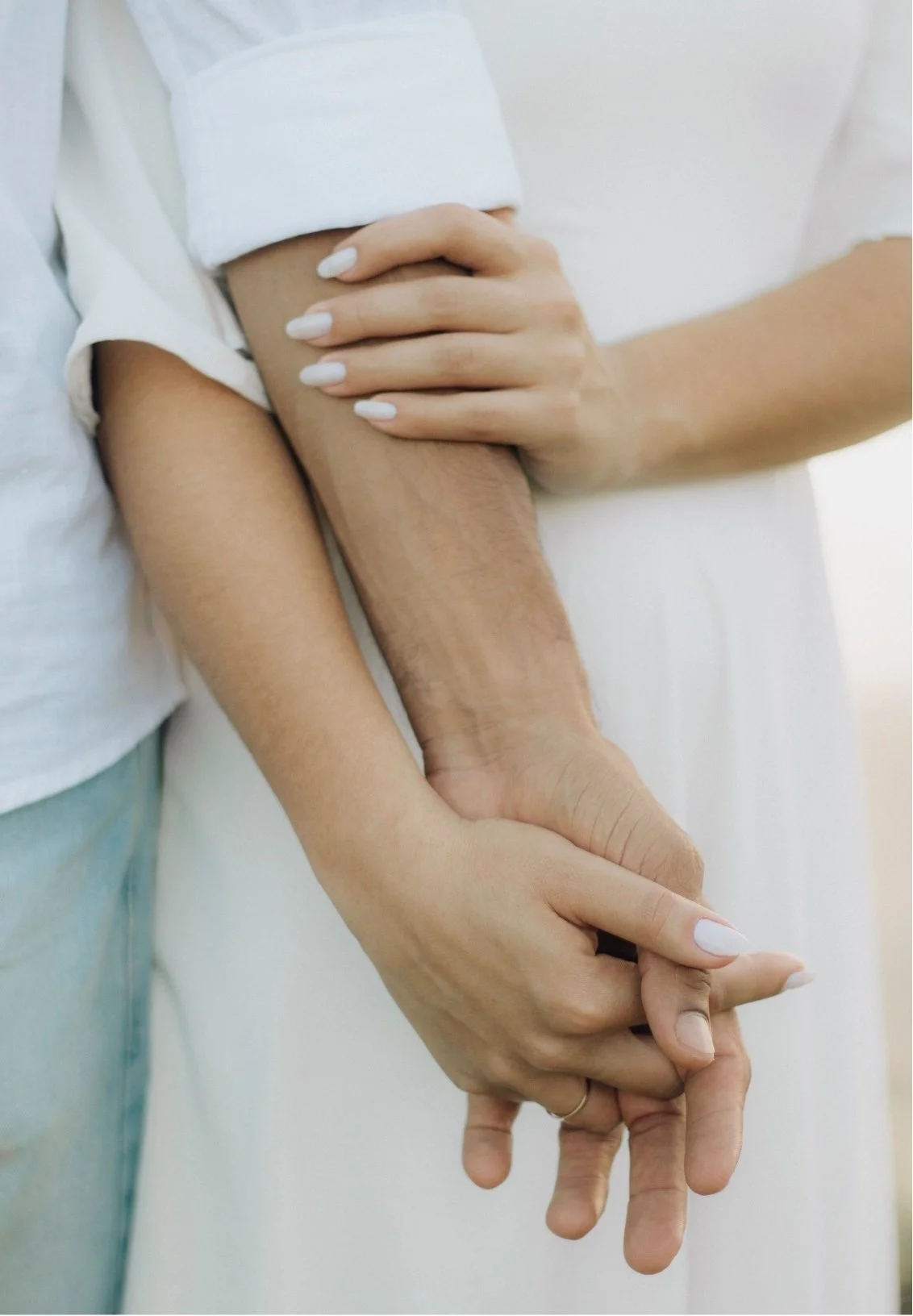 Two people holding hands, one with a darker skin tone and the other with a lighter skin tone, lying on a white surface.
