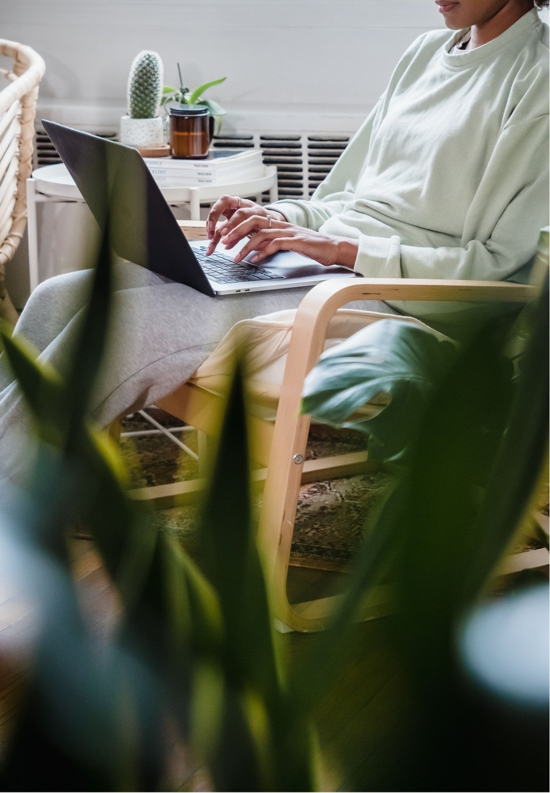 A person sitting in a wooden chair using a laptop, with indoor plants and a stack of magazines or books in the background.