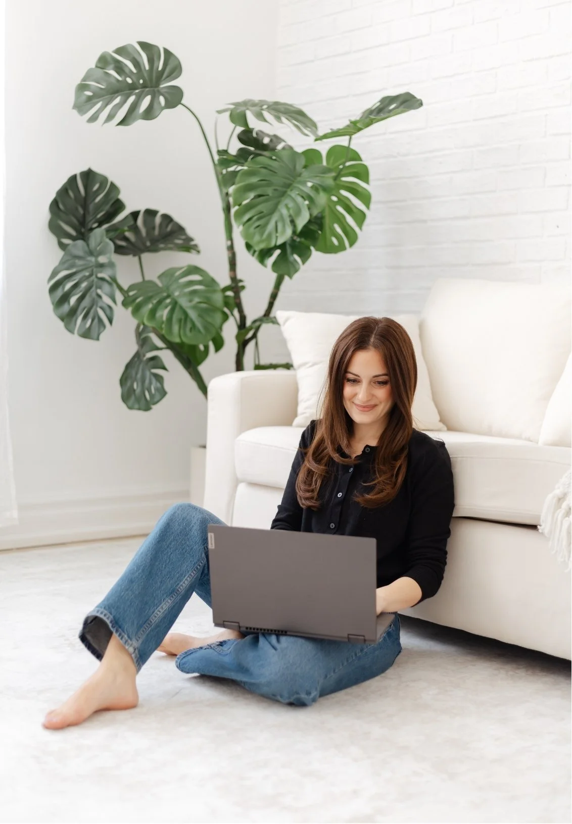 A woman sitting on the floor with a laptop on her lap, smiling, in a bright, minimalist living room with a white sofa, cushions, a large green monstera plant, and a white brick wall.