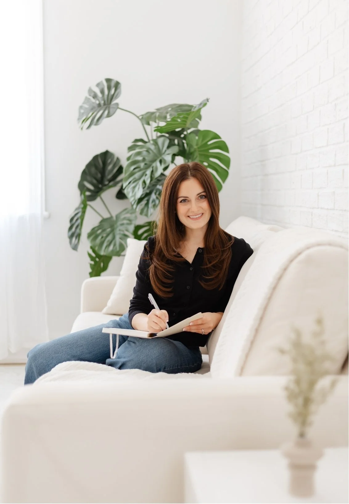 Samantha Mirarchi, therapist in Richmond Hill, sitting on a white couch with a notepad and pen, smiling, in a bright room with a large green plant in the background.