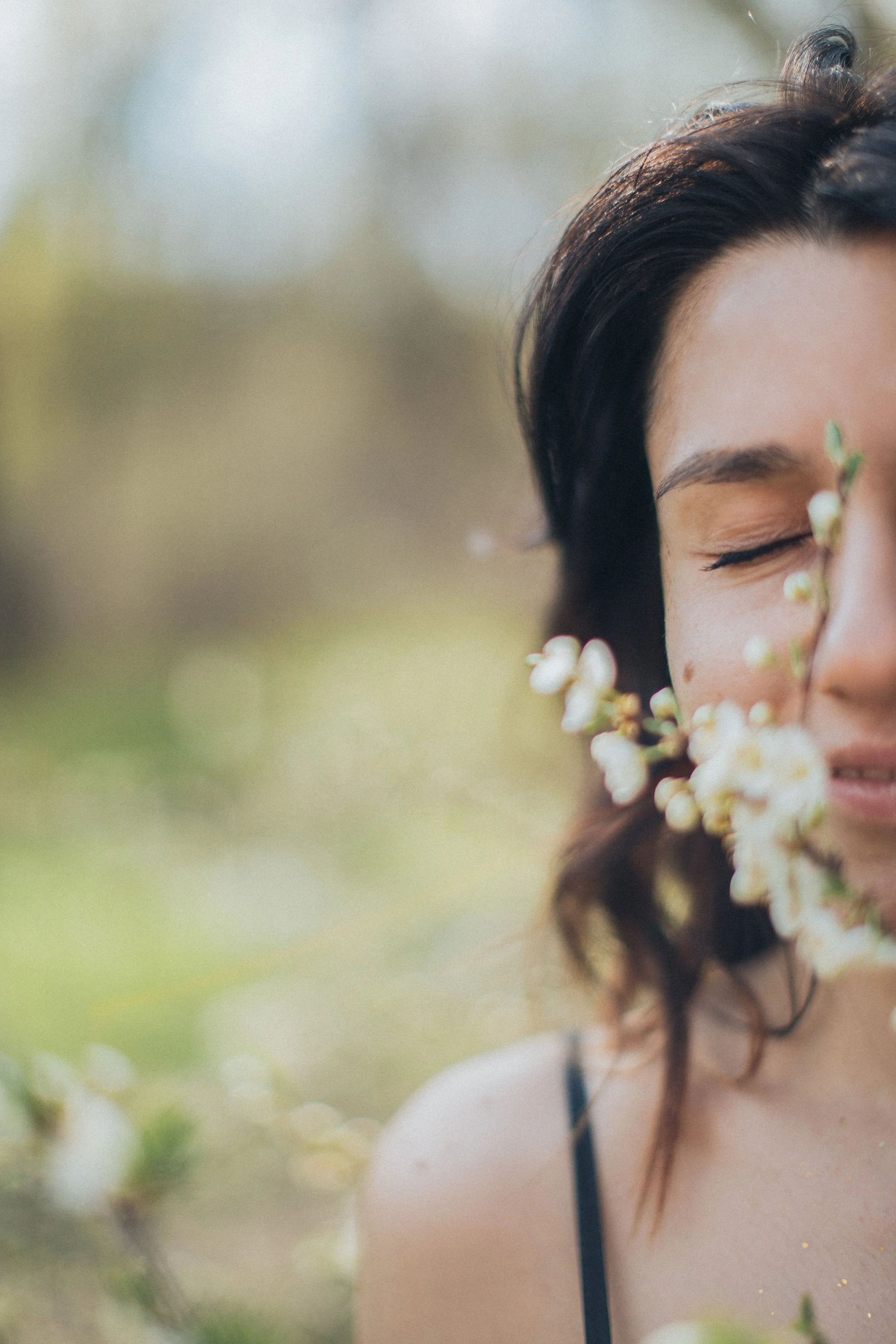 A close-up of a woman with dark hair and closed eyes, standing outdoors with blurred greenery in the background, holding white flowers near her face.