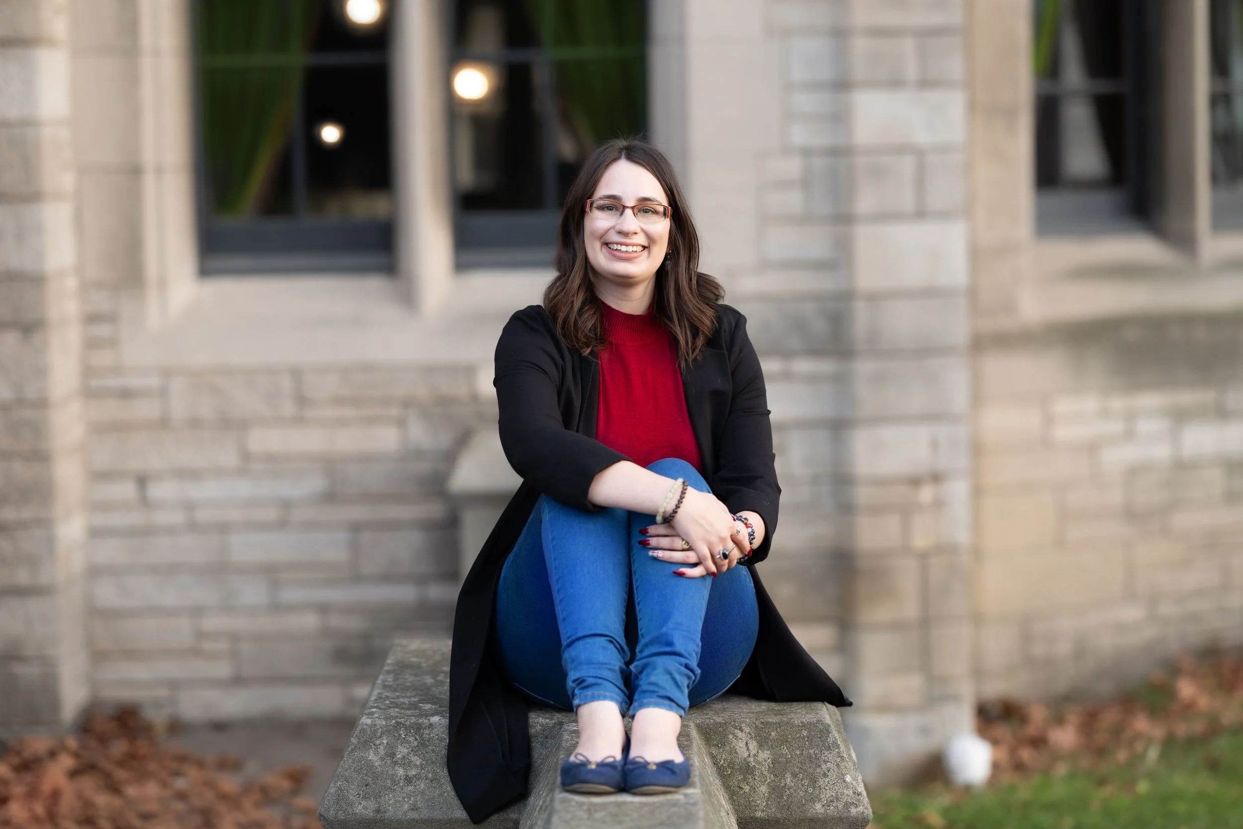 Hillary Bowdridge, psychotherapist in Richmond Hill, ON, sitting on a concrete ledge outside a stone building with large windows, smiling at the camera, wearing glasses, a red top, black jacket, and blue jeans.