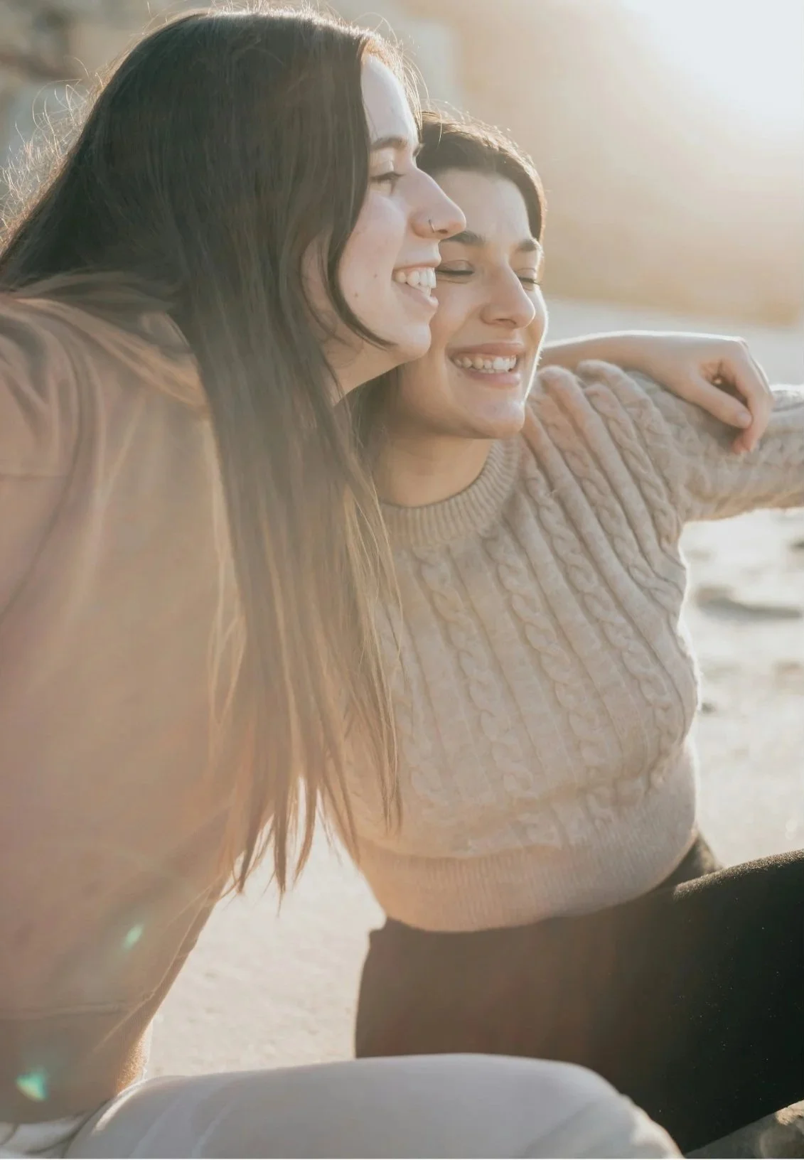 Two women smiling with eyes closed, taking a selfie outdoors during sunset, one has long dark hair and the other has short dark hair, wearing a beige knitted sweater.