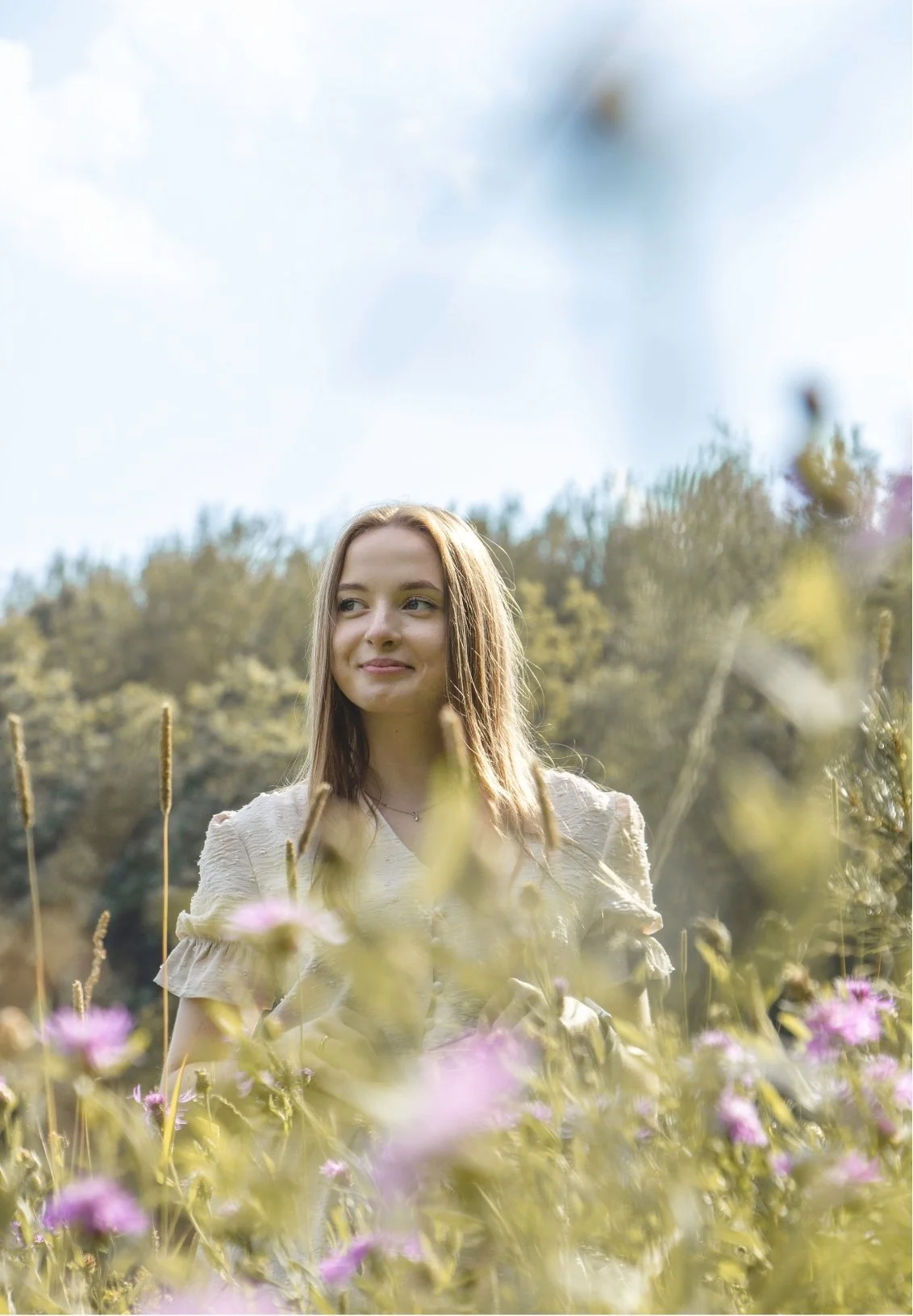 A young woman with long brown hair standing in a field of wildflowers, smiling softly, with a backdrop of trees and a partly cloudy sky.
