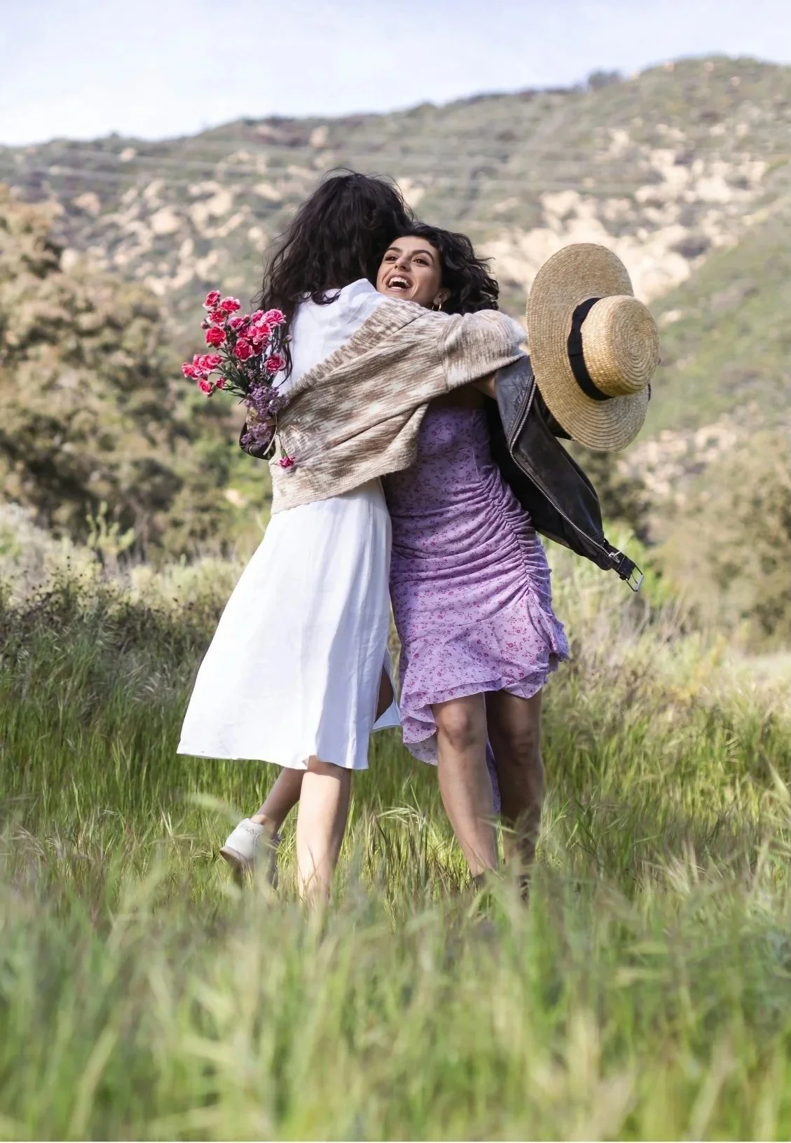 Two women hugging outdoors in a grassy field with hills in the background. One woman holds a straw hat, and the other has a bouquet of pink flowers on her back. They are smiling and appear happy.