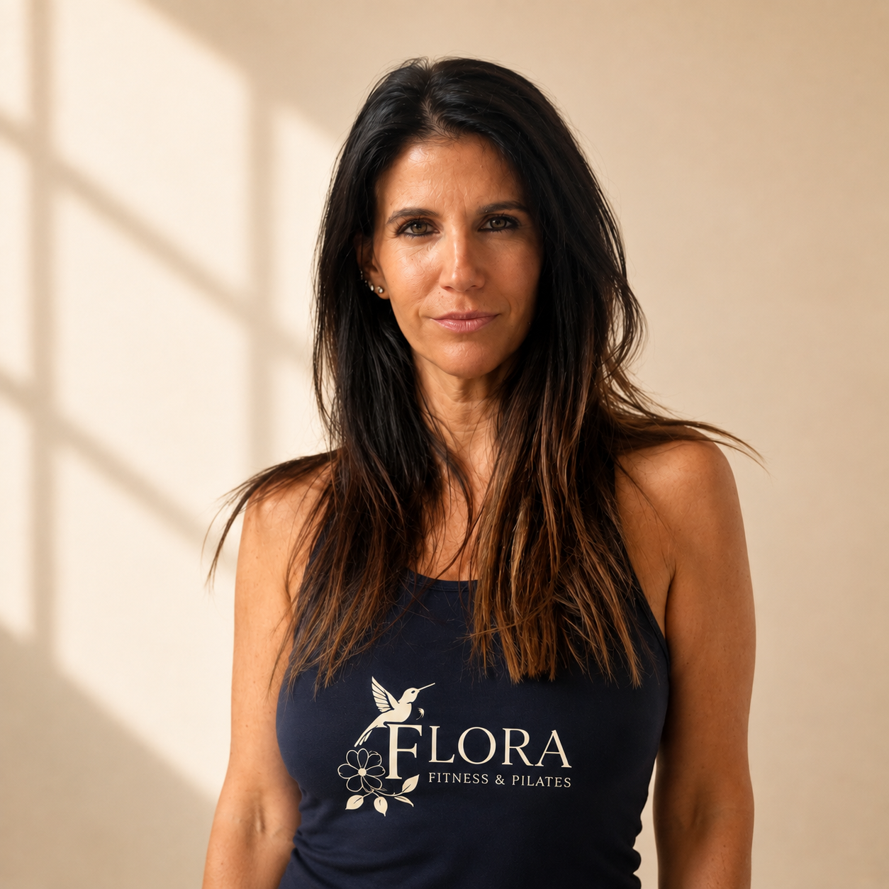 A woman with long, dark hair wearing a navy tank top with a logo that says "Flora Fitness & Pilates" with a hummingbird and flower design.
