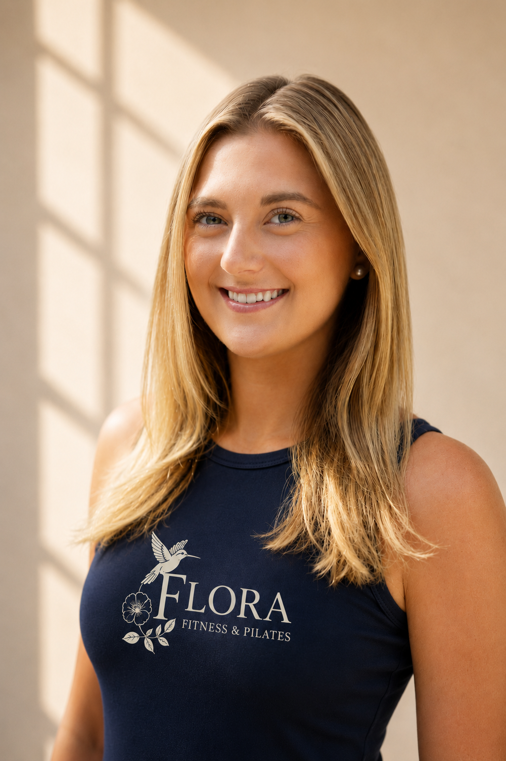 A woman with long blonde hair smiling, wearing a navy sleeveless shirt with 'Flora Fitness & Pilates' and a hummingbird logo, standing near a sunlit wall.