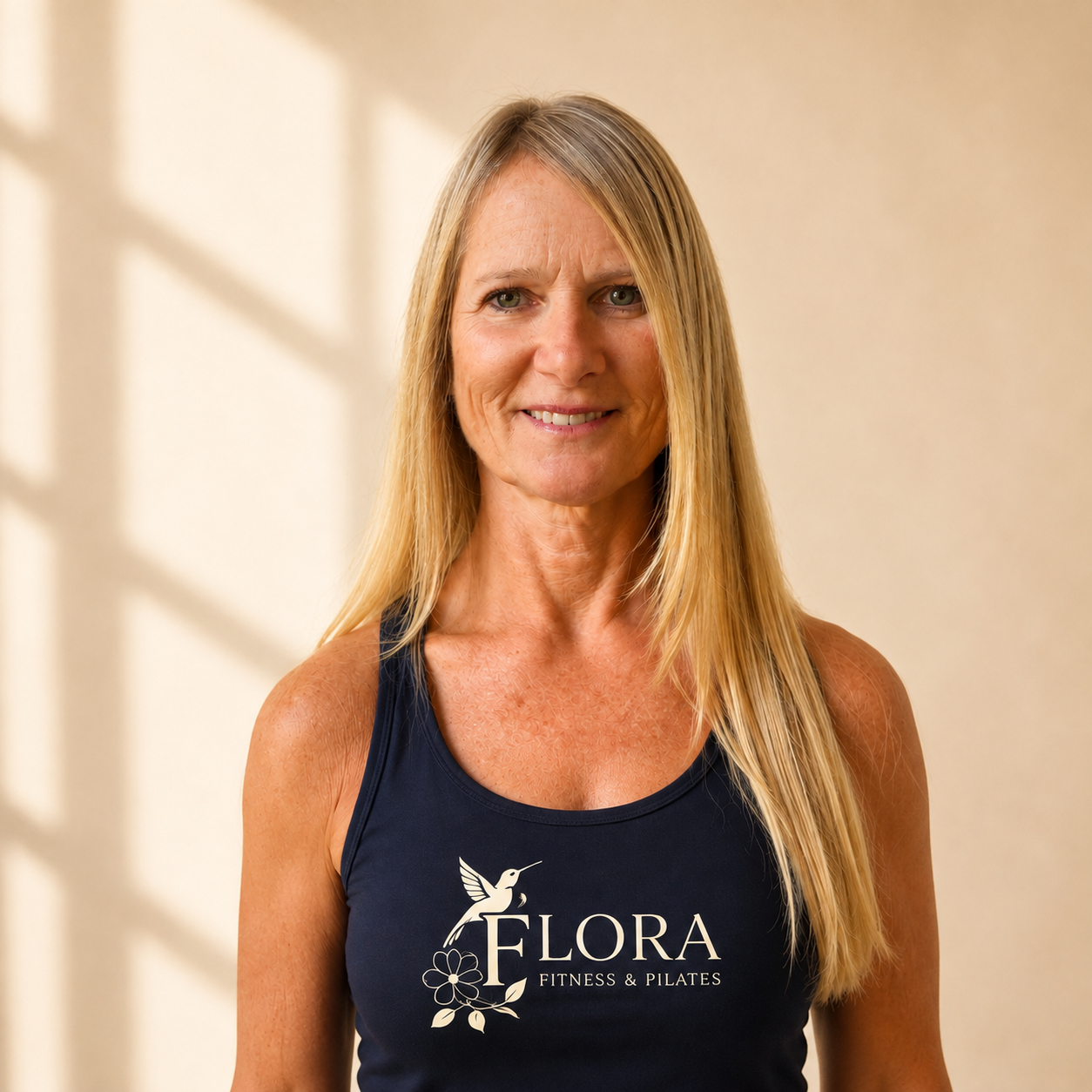 A woman with long blonde hair smiling, wearing a navy tank top with a hummingbird and flower logo and the text "Flora Fitness & Pilates" on it, standing indoors with a beige wall and shadows in the background.