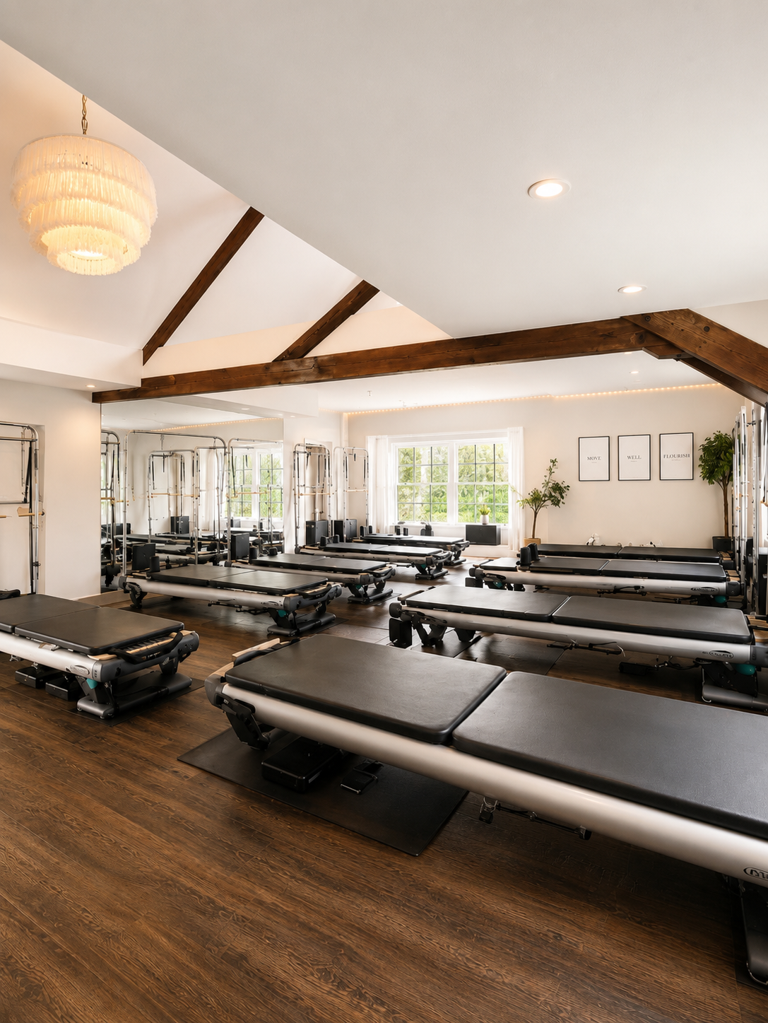 A spacious Pilates studio with multiple reformer machines arranged in rows near a large window with greenery outside, decorated with framed minimalist art on the wall.
