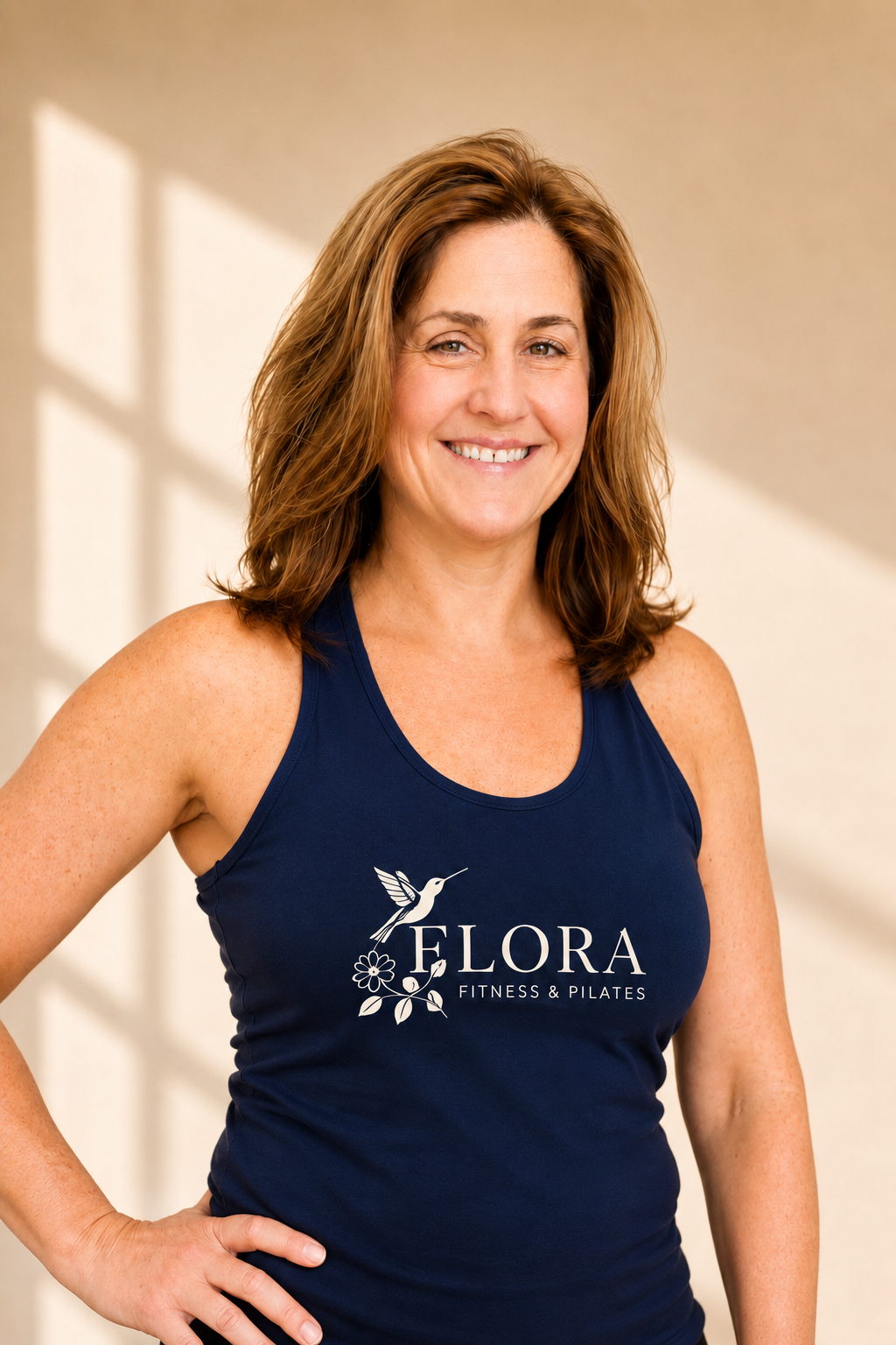 Woman smiling, wearing a navy tank top with the logo 'Flora Fitness & Pilates' featuring a hummingbird and floral design.
