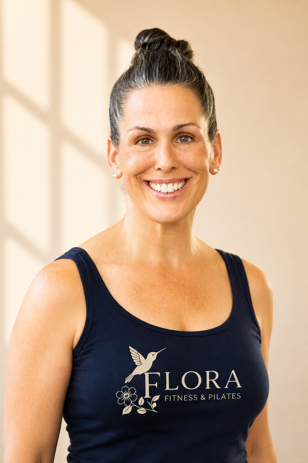 Smiling woman with dark hair in a bun, wearing pearl earrings and a navy tank top with 'FLORA FITNESS & PILATES' logo.