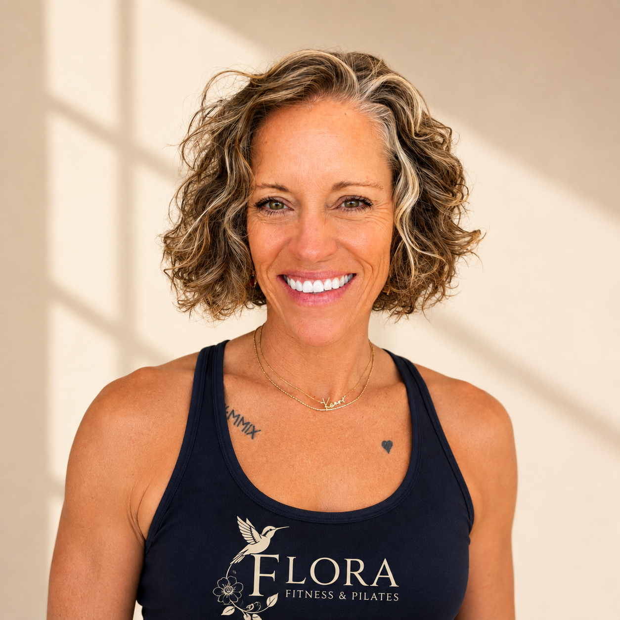 A smiling woman with short, curly gray hair wearing a navy blue tank top that says 'FLORA FITNESS & PILATES'. She is wearing two dainty necklaces, one with a name and the other plain. She has small earrings and two tattoos, one reading 'XIMMI' on her left collarbone and a small heart on her chest. The background has soft natural light with a shadow pattern.