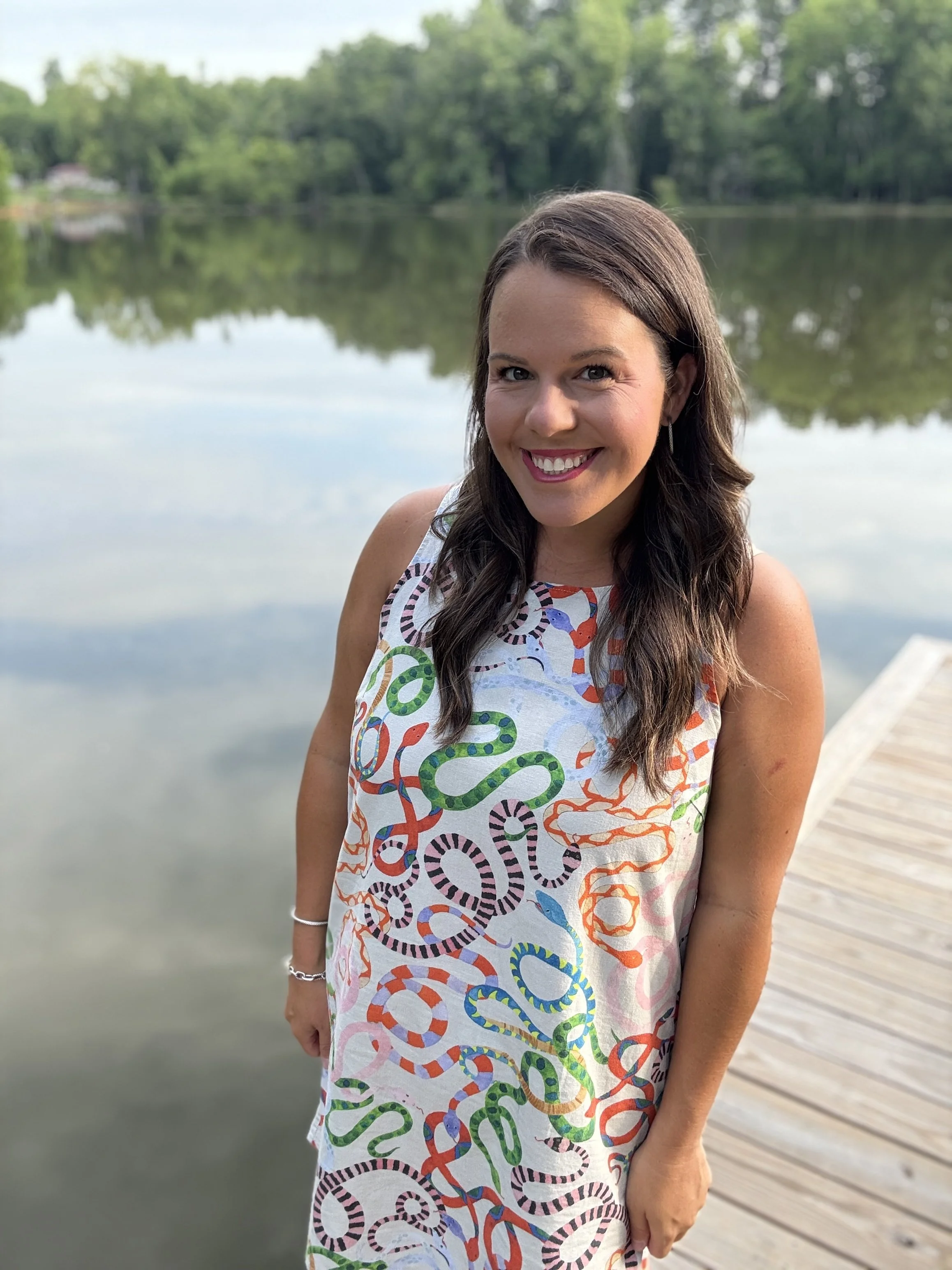 A smiling woman with long dark hair, wearing a white dress with colorful snakes and loops pattern, standing on a wooden dock by a calm lake with green trees in the background.