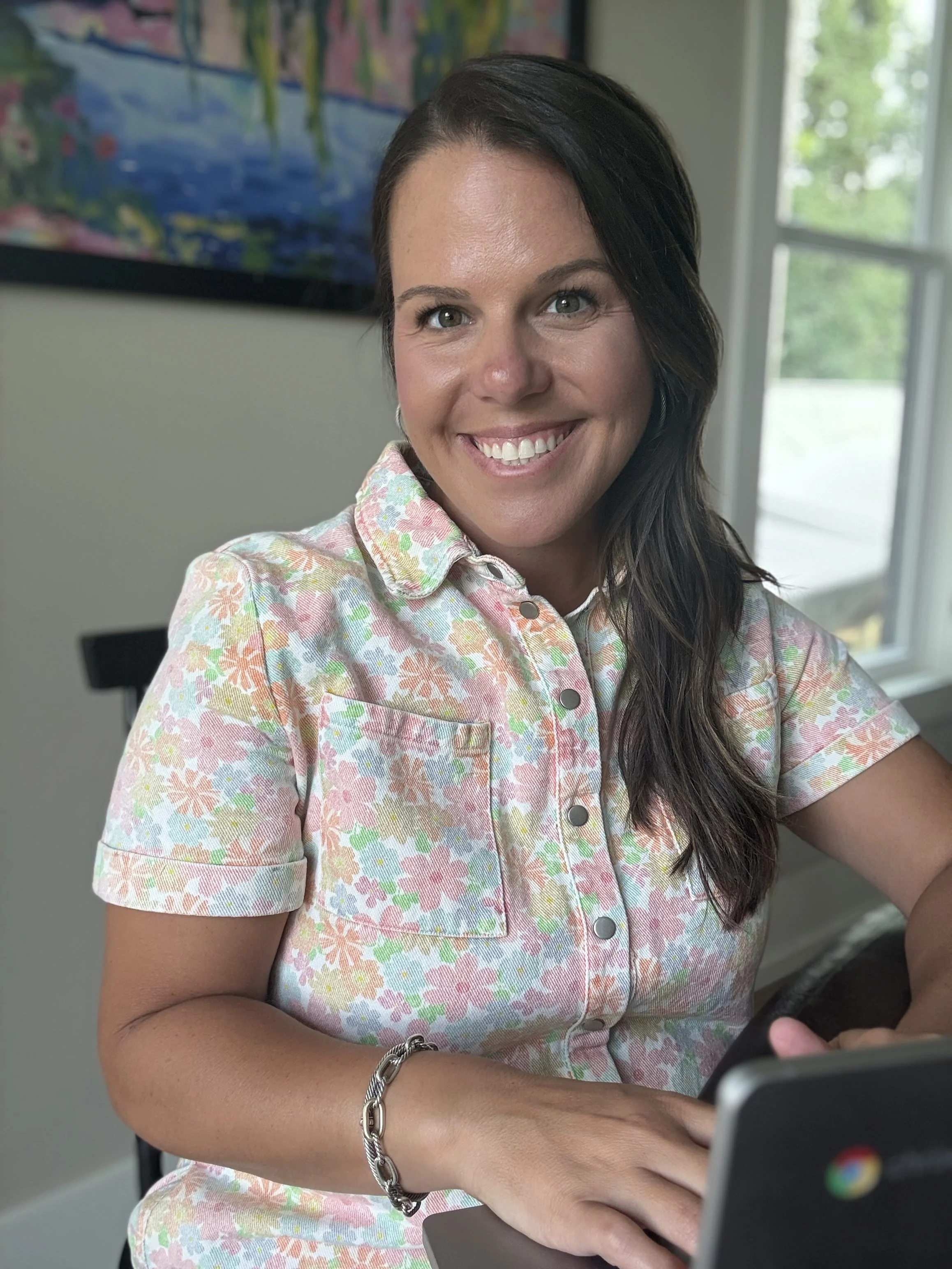 A woman with long dark hair and light skin, smiling and looking at her phone, wearing a short-sleeved pastel floral shirt and a silver chain bracelet, with a colorful painting and window in the background.