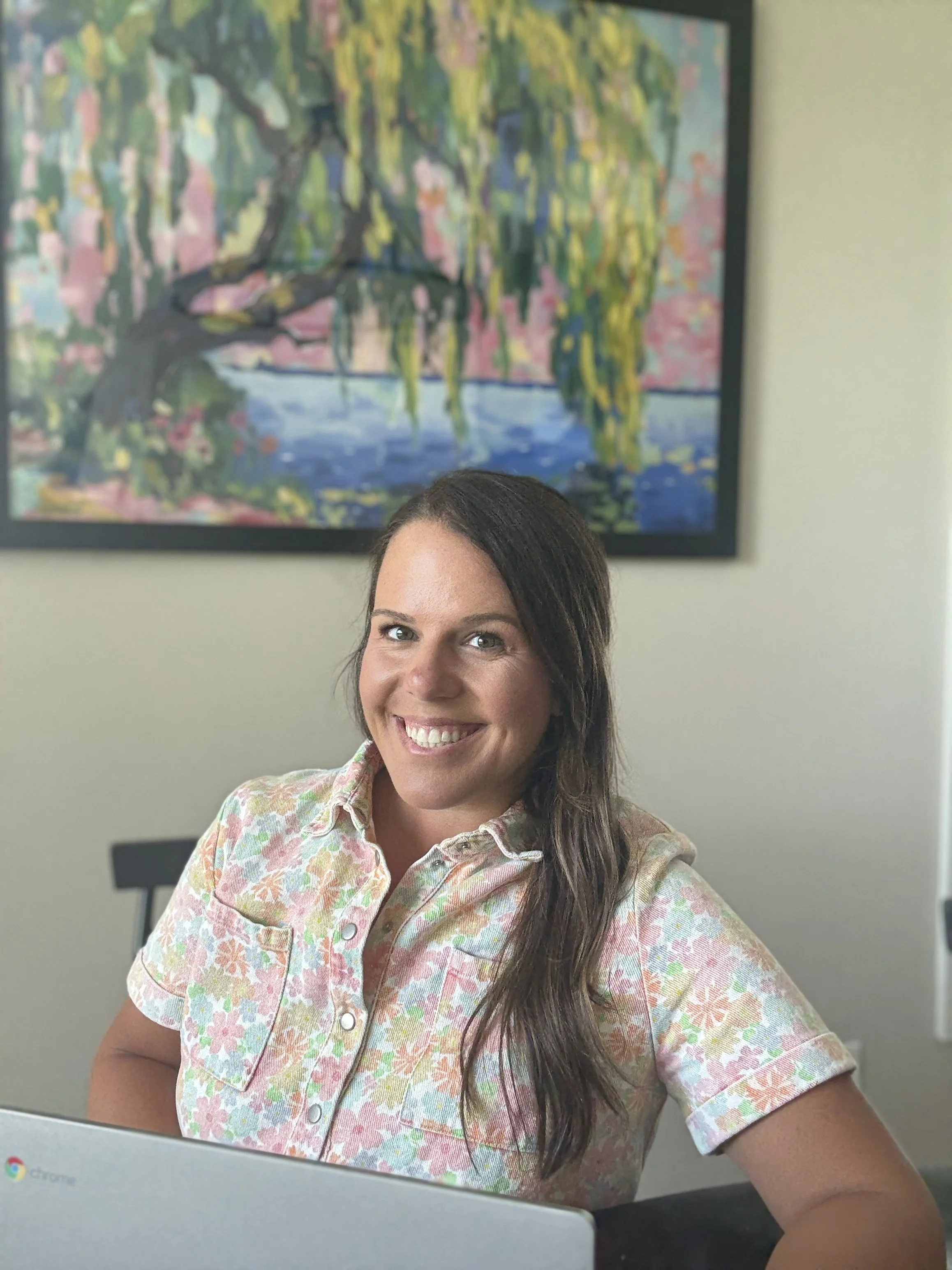 A woman with long dark hair and a bright smile sitting in front of a laptop and smiling at the camera. She is wearing a pastel floral shirt. Behind her is a framed painting of a willow tree over a pond with a pink and green background.