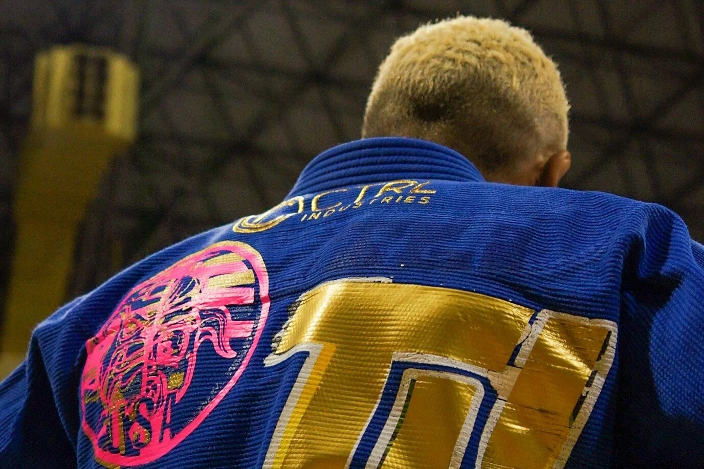 Close-up of a person wearing a blue judo gi with patches, seen from behind, with a yellow and black background.