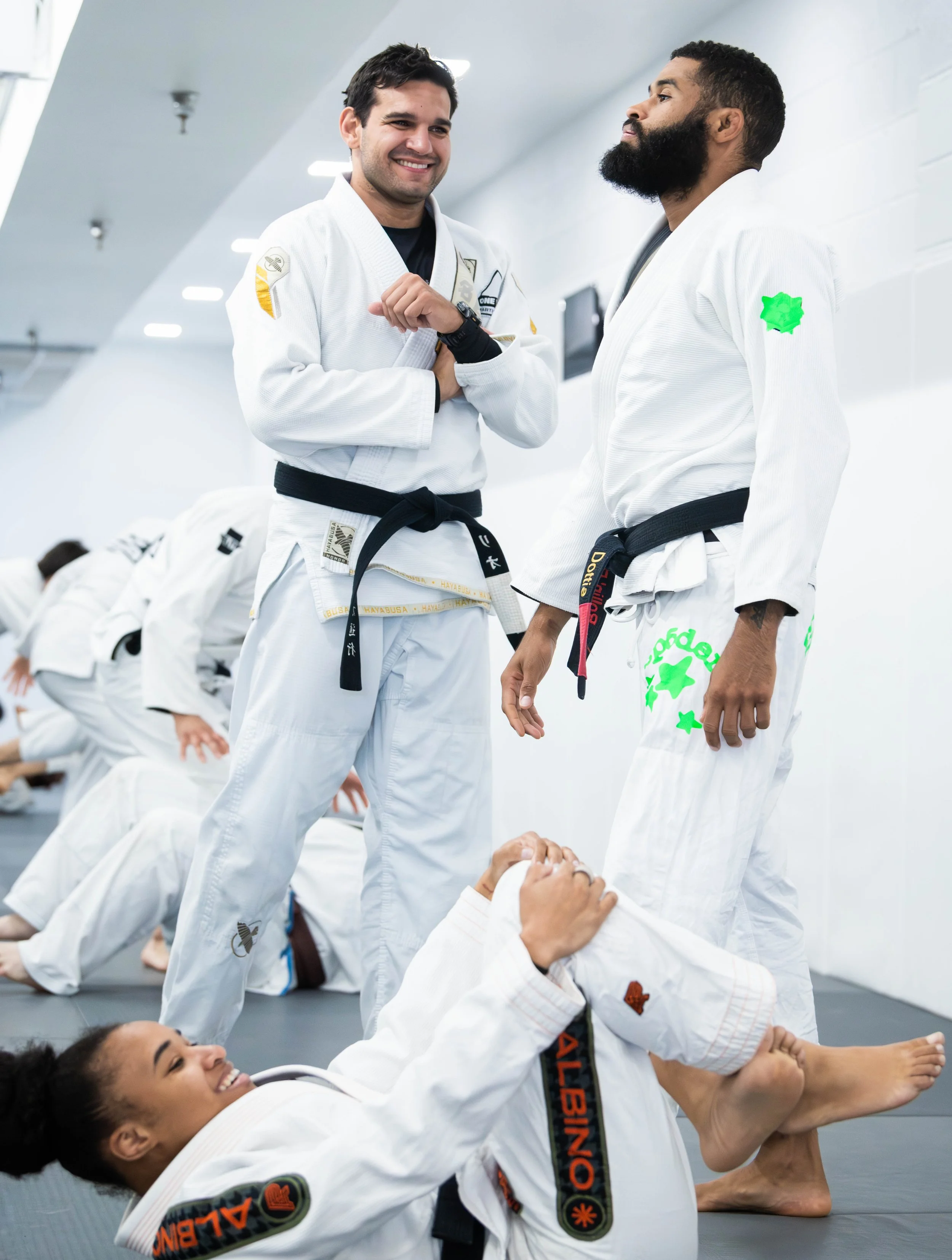 Two men in white Brazilian Jiu-Jitsu gis, with black belts, standing and smiling in a martial arts gym. One man has a beard, and the other has short dark hair. They are engaged in a conversation, and others are practicing on the floor behind them.