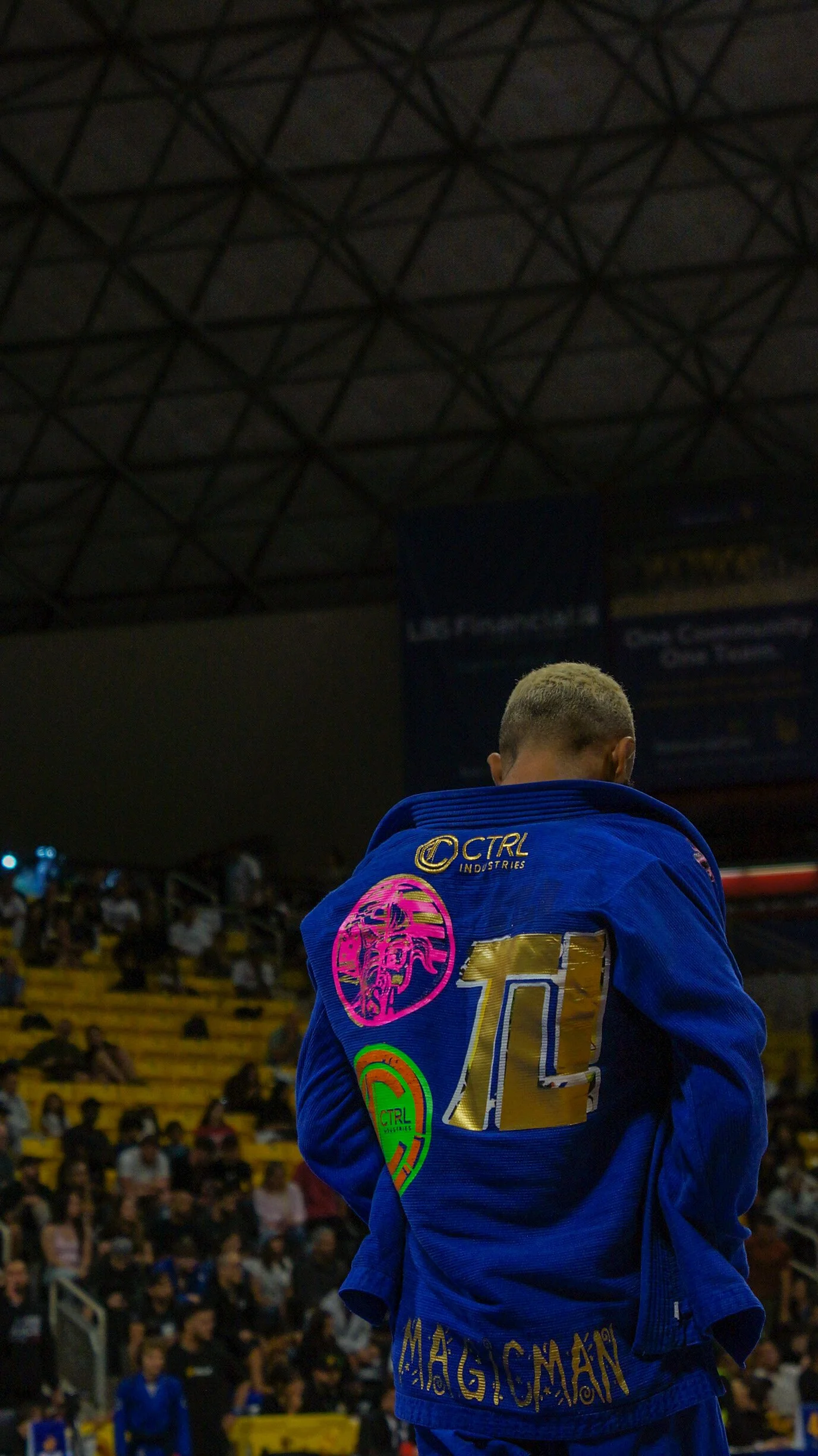 Back of a Brazilian Jiu-Jitsu competitor wearing a blue gi with patches, standing in a crowded gymnasium with an audience and banners in the background.