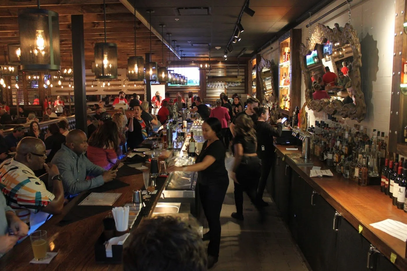 Inside a busy bar with patrons seated and bartender preparing drinks, warm lighting, wooden decor, and bottles of alcohol on the counter.