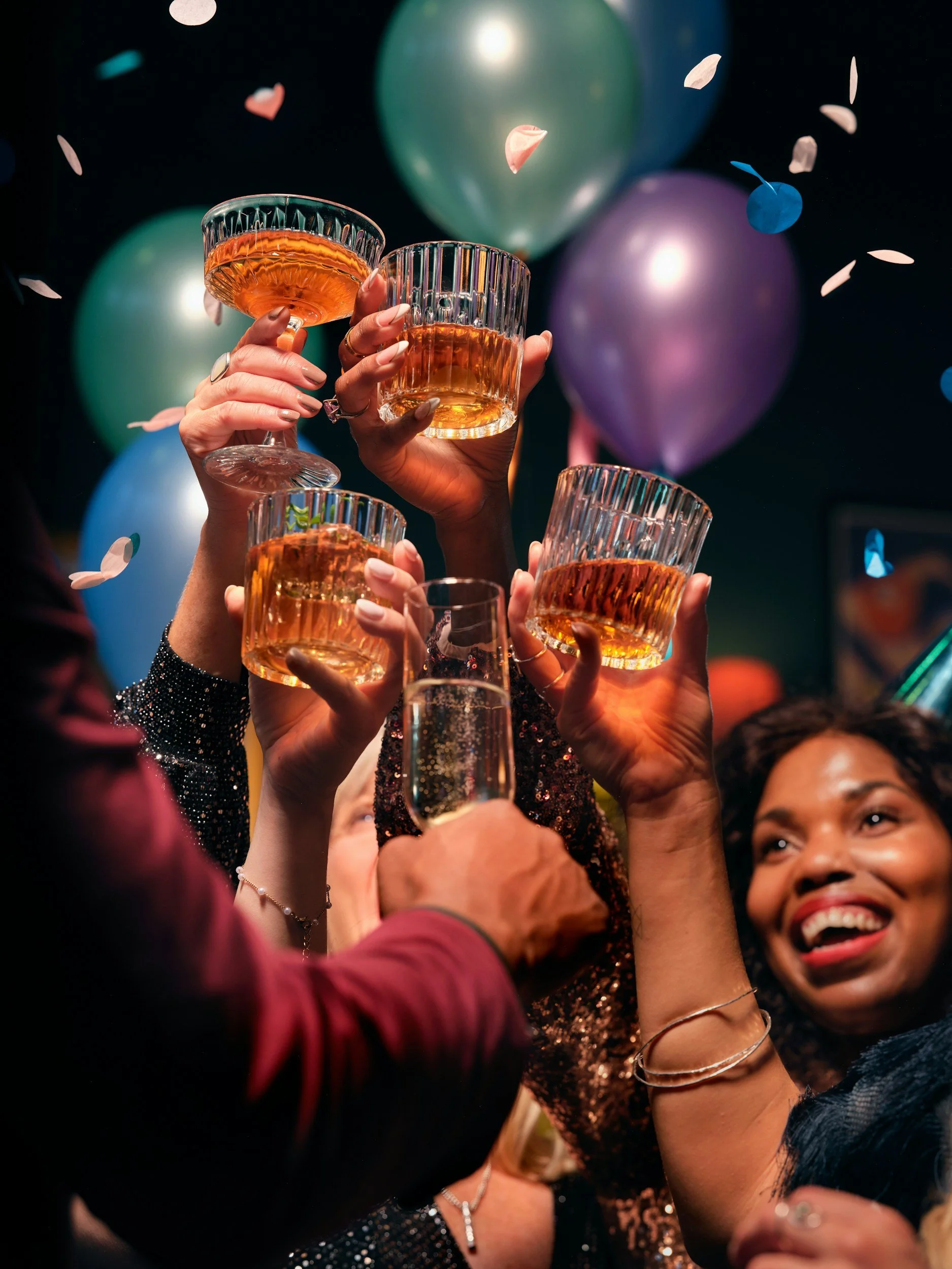 People celebrating with raised glasses of champagne or wine amidst colorful balloons and confetti, smiling and enjoying a festive occasion.