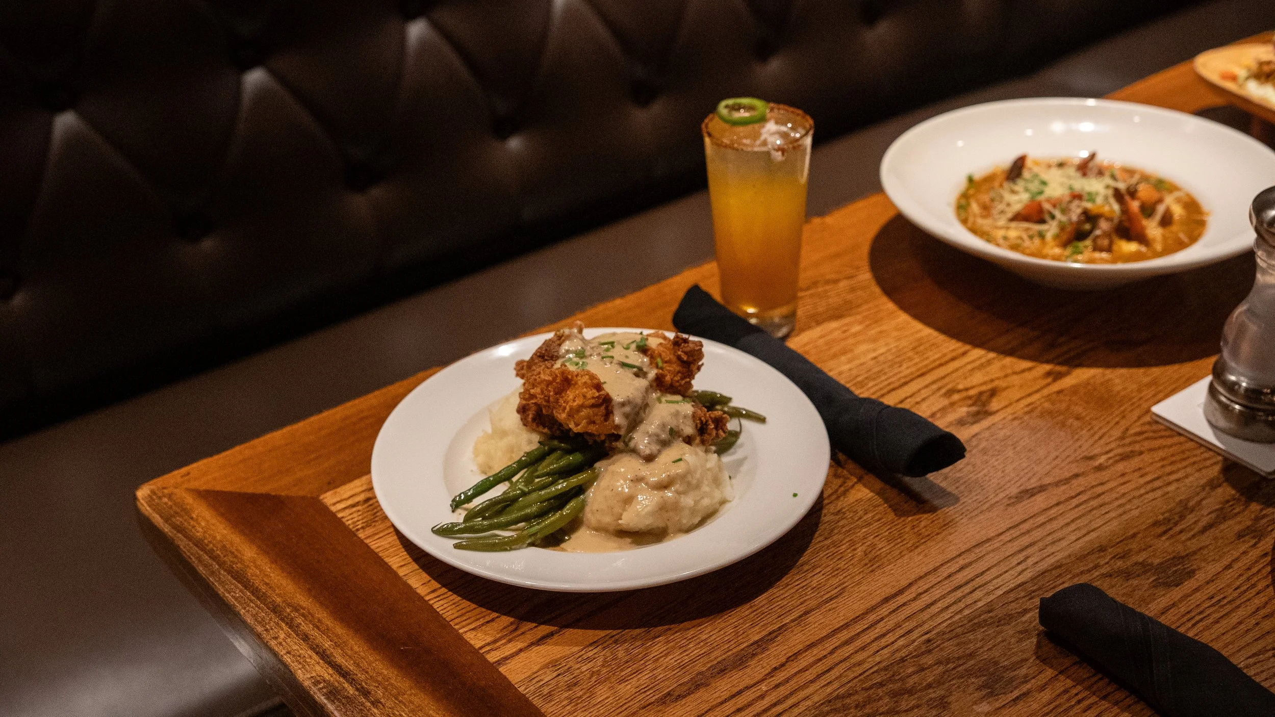 Wooden table with a plate of fried chicken, green beans, mashed potatoes with gravy, a bowl of pasta with sauce and vegetables, a tall glass of yellow beverage with a lime, salt and pepper shakers, and a napkin with cutlery.