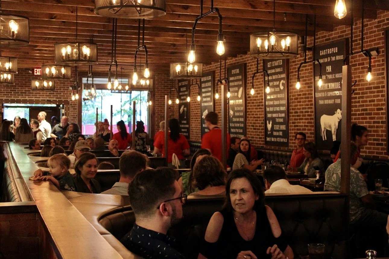 A busy restaurant with brick walls, hanging Edison bulb lights, and several patrons seated at tables and booths, with some servers attending to customers.