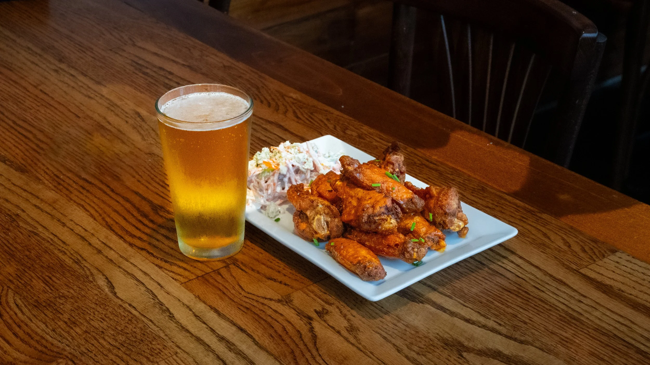 A white rectangular plate with fried chicken wings, potato salad, and green garnish, a tall glass of beer on a wooden table.