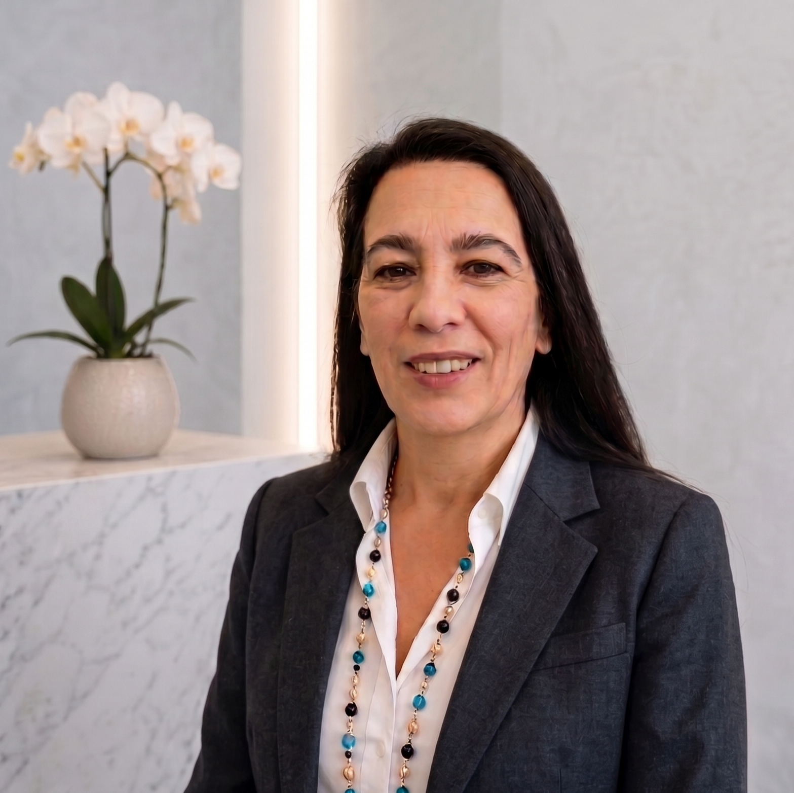 A middle-aged woman with dark hair, wearing a gray blazer, white shirt, and a multi-colored beaded necklace, smiling at the camera in an office or lobby setting with a white orchid in a pot in the background.