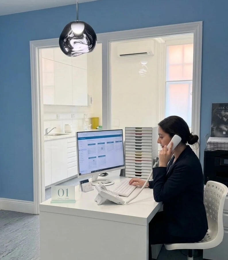 Woman working at a white desk in an office, talking on a landline phone, with a computer monitor, keyboard, and filing trays, near a window and a blue wall.