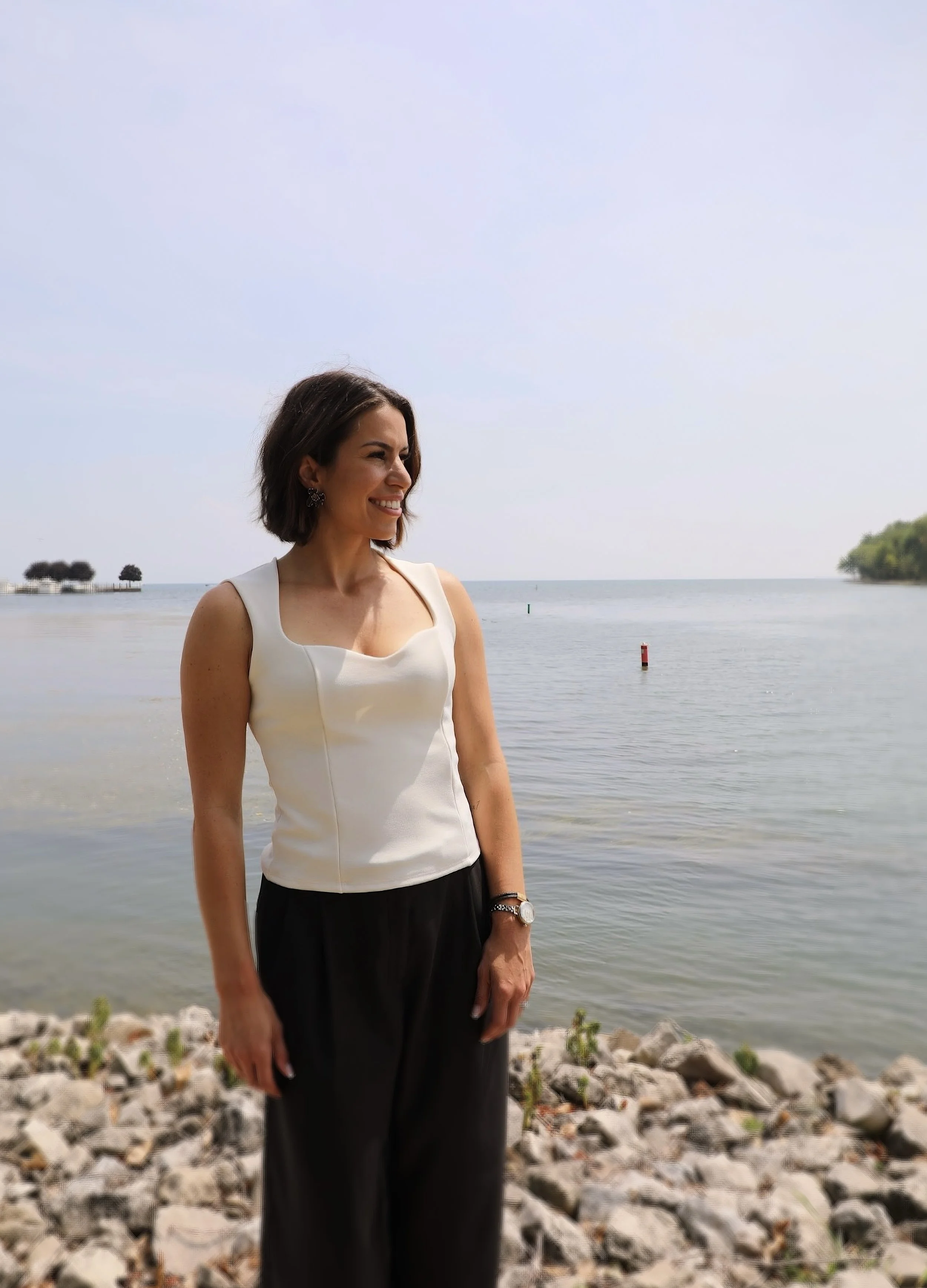 A woman with short dark hair smiling and standing outdoors by a body of water. She is wearing a white sleeveless top, black pants, a watch, and earrings, with rocks and greenery in the background.