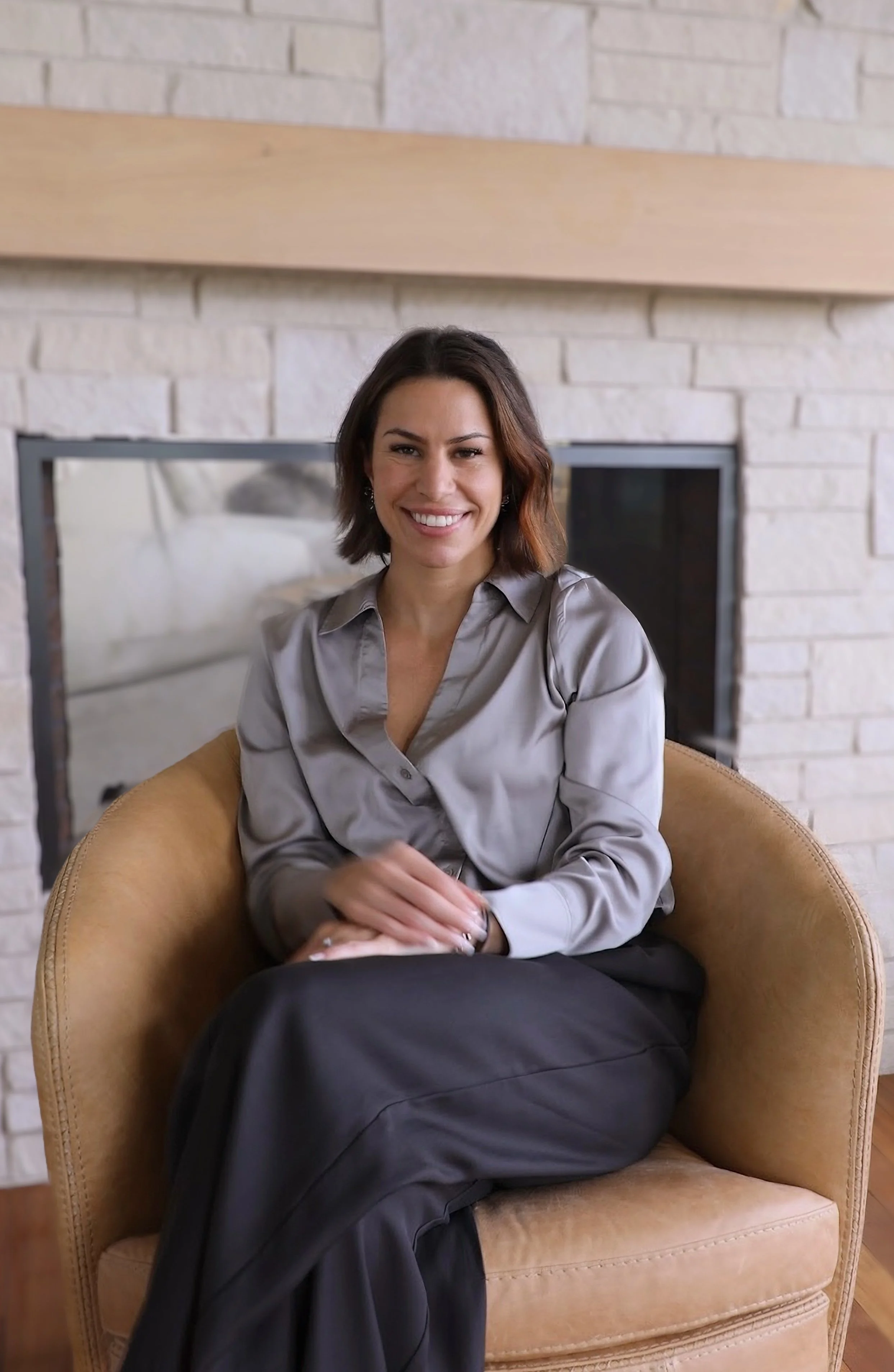 A woman with shoulder-length dark hair and a gray silk shirt, sitting comfortably on a tan armchair in a living room with a brick fireplace in the background, smiling at the camera.