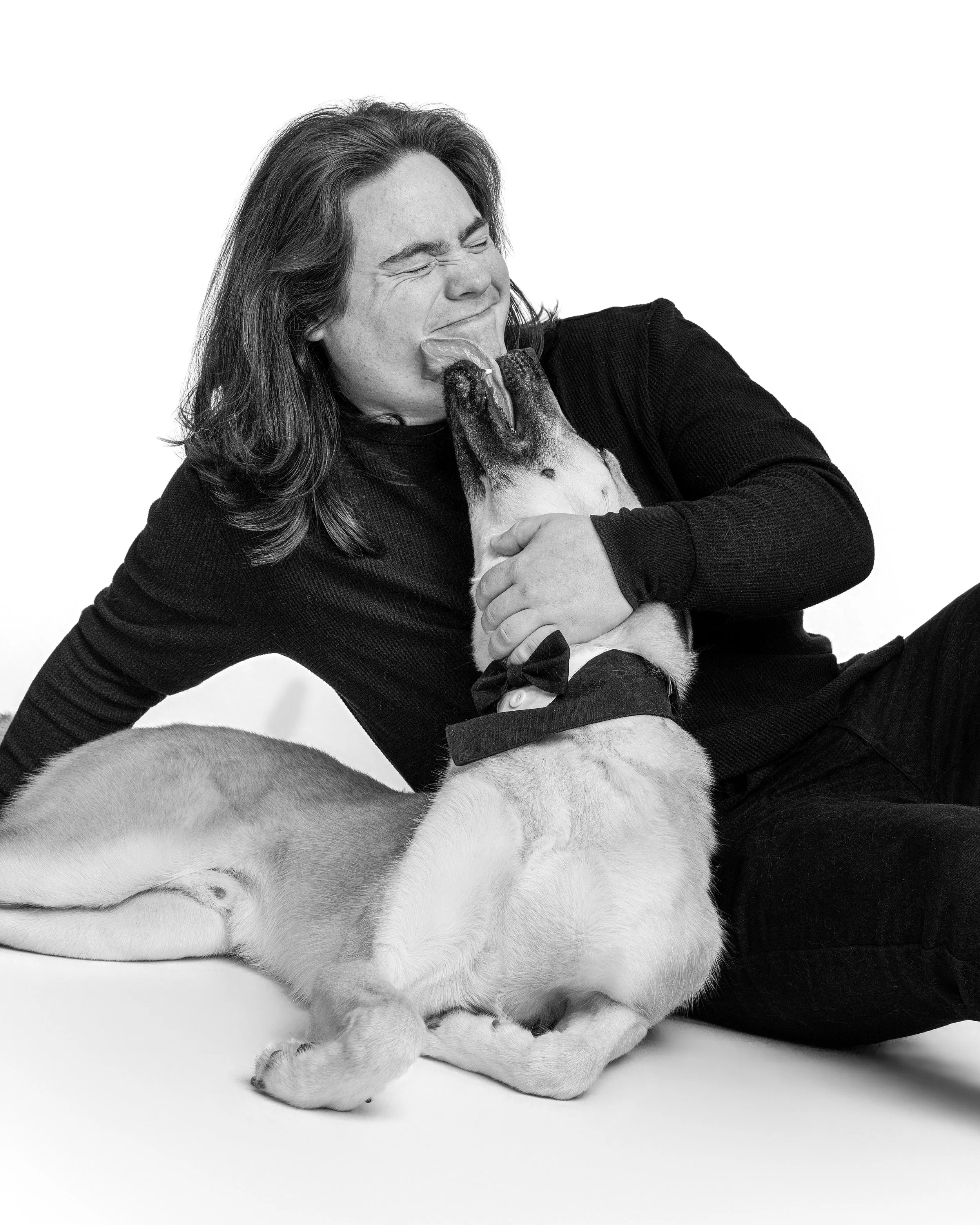 A man with long hair squeezes his eyes shut and laughs as his light-coated dog, wearing a black bow tie, licks his face in a black and white studio portrait.
