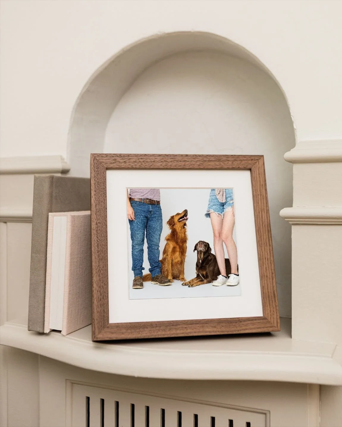 Walnut frame with a white mat on a mantel shelf holding a studio portrait of a golden retriever and a chocolate Lab looking up at their people, shown from the knees down