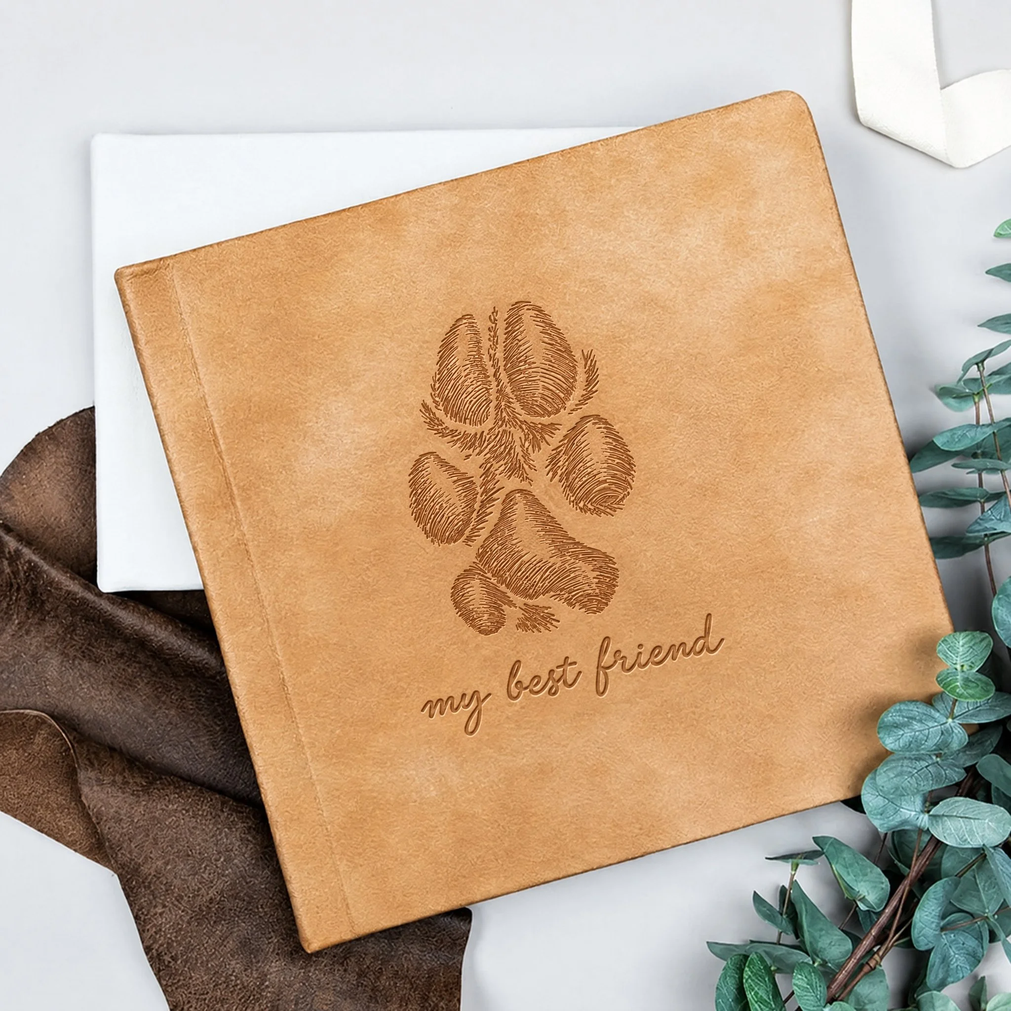 Tan suede leather fine art album with a paw print embossed on the cover above the words my best friend, resting on brown leather with eucalyptus leaves