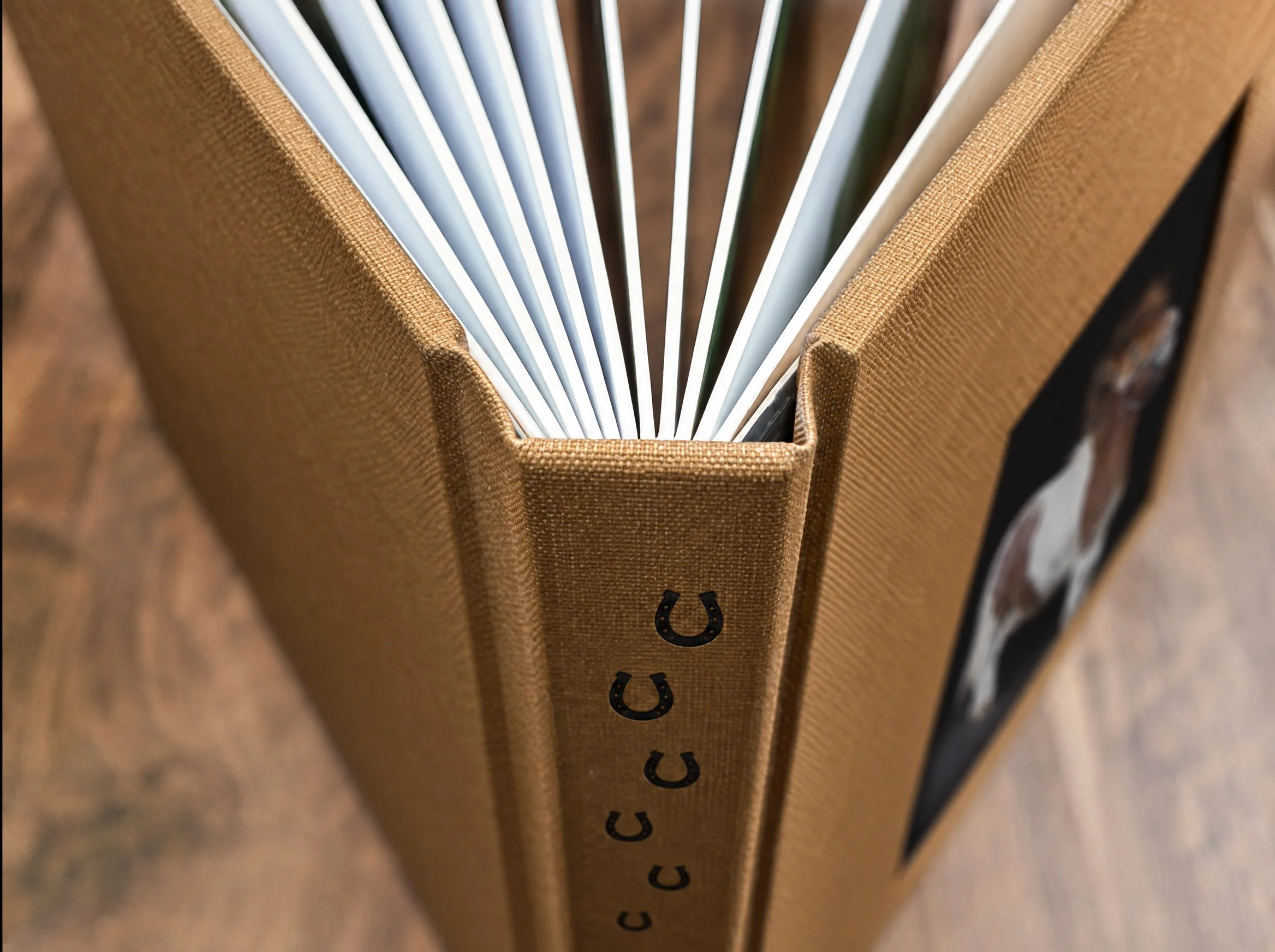 Tan linen horse portrait album standing on its spine with horseshoe icons stamped down the spine and pages fanned open at the top