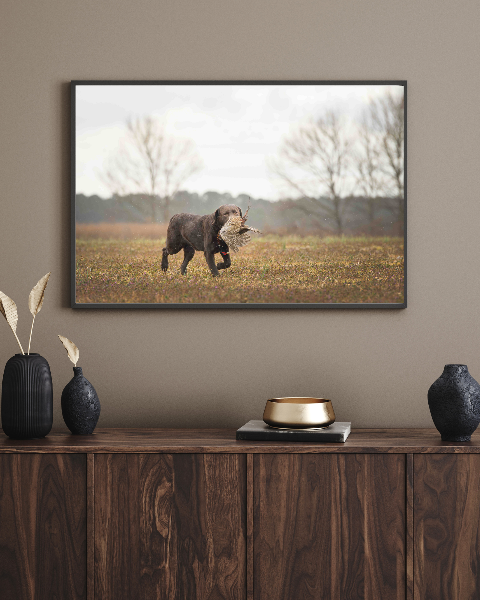 Framed photograph of a brown hunting dog carrying a pheasant in a field above a wooden sideboard with decorative vases.