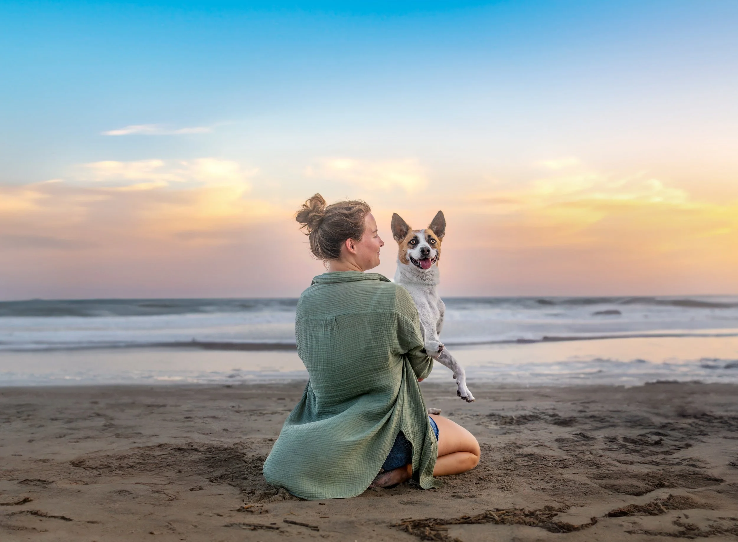 A woman in a sage green shirt kneels in the sand holding her red and white dog, who grins at the camera as the sky turns pink over the ocean.