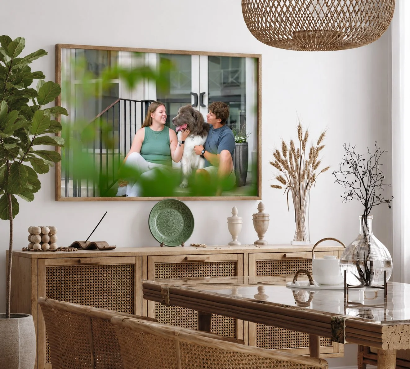 A framed portrait of a couple in sage green and soft blue with their Newfoundland, hung in a dining room with rattan, warm wood, and green accents.
