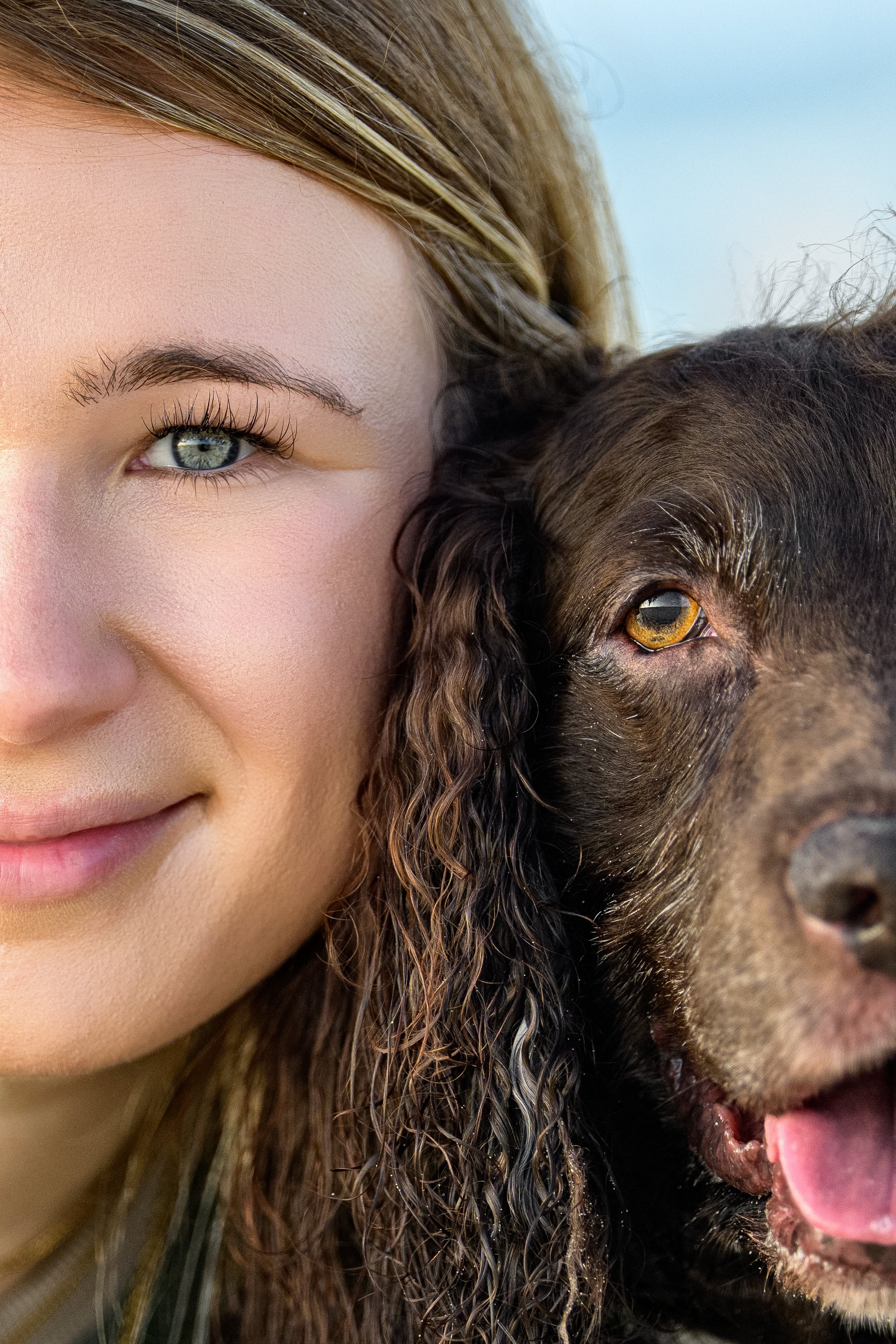 A young woman and her Boykin Spaniel pressed cheek to cheek in a close crop, her blue green eye and his amber eye both looking at the camera.