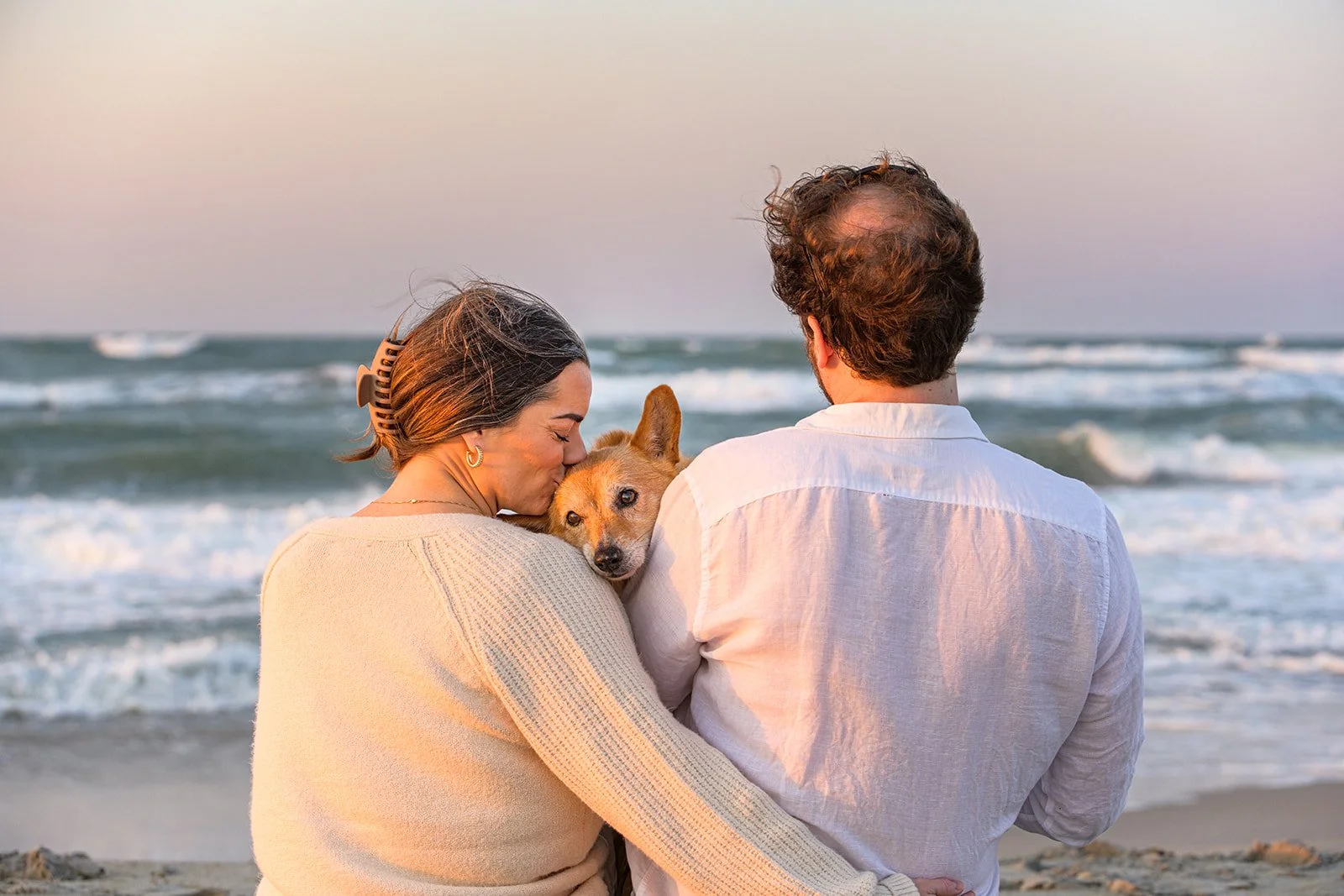 A woman with her hair in a claw clip kisses her corgi on a windy beach as waves roll in behind them.