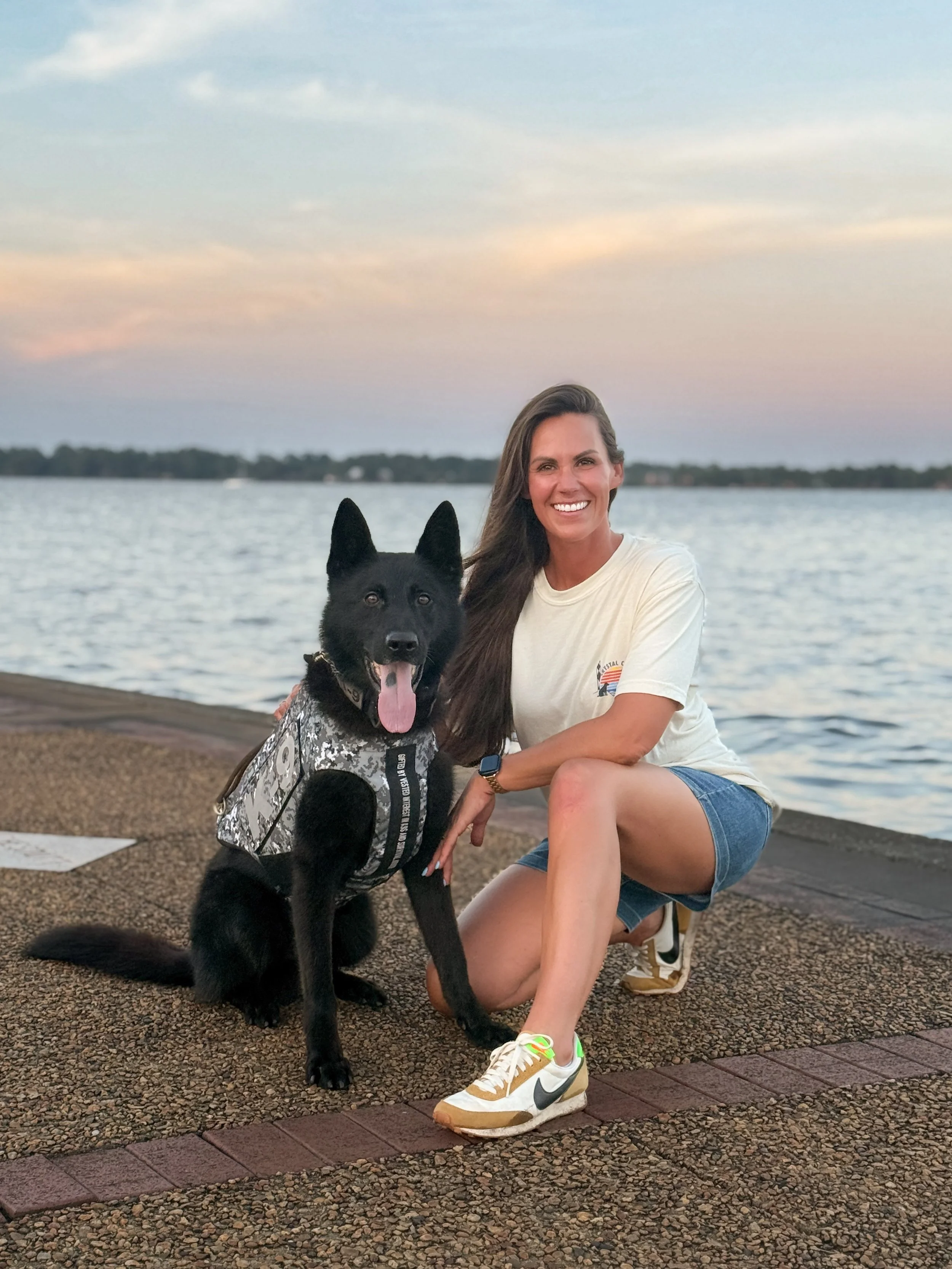 Smiling woman kneeling beside a black German Shepherd wearing a patterned service vest near a lake at sunset
