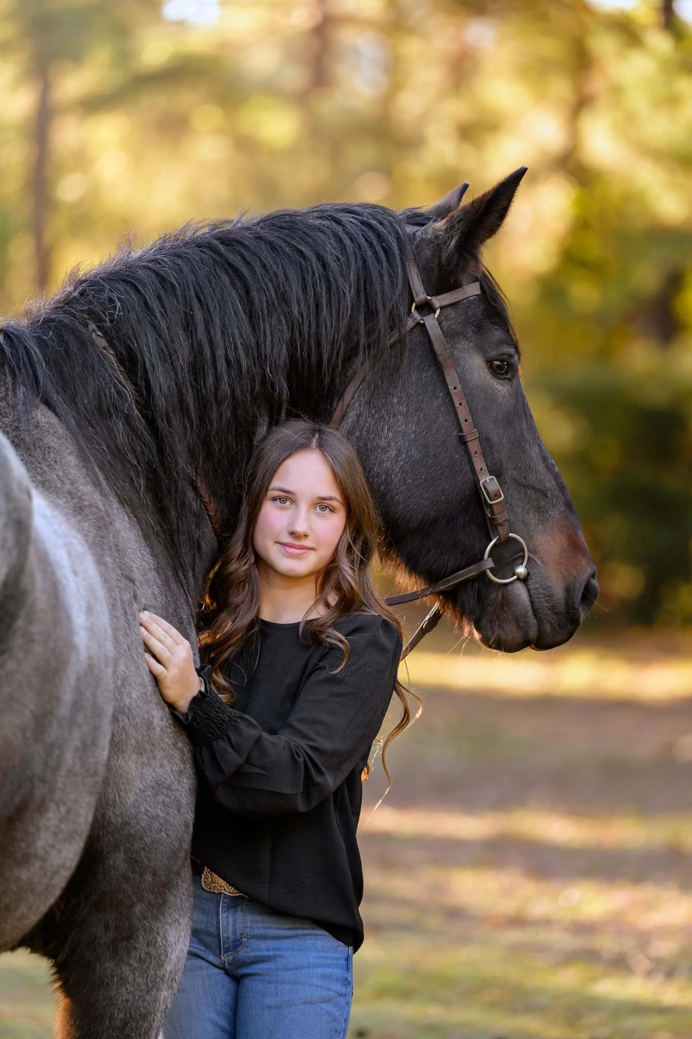 Young woman standing beside a large black horse wearing a bridle in a sunlit forest