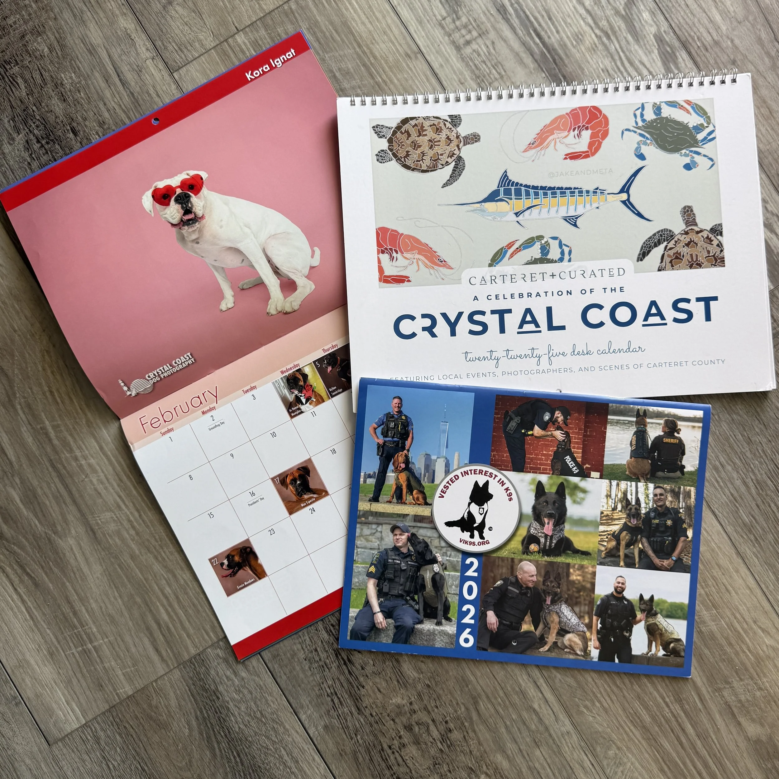 Calendars spread across a wood floor, including a dog photography calendar, a Crystal Coast wildlife calendar, and a police K-9 calendar.