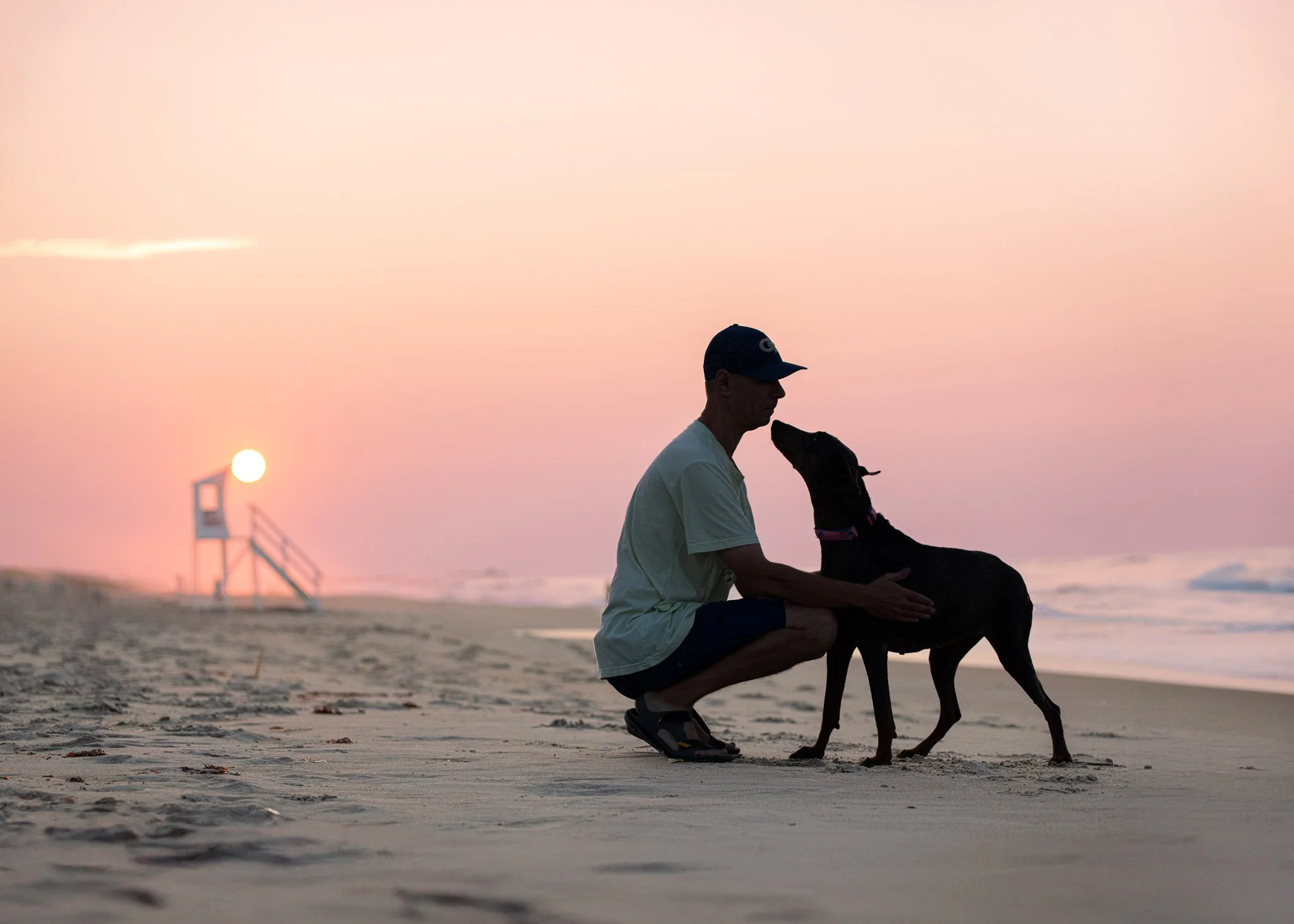 A man kneels nose to nose with his senior Doberman on the beach at sunrise, the sun rising beside a lifeguard stand.